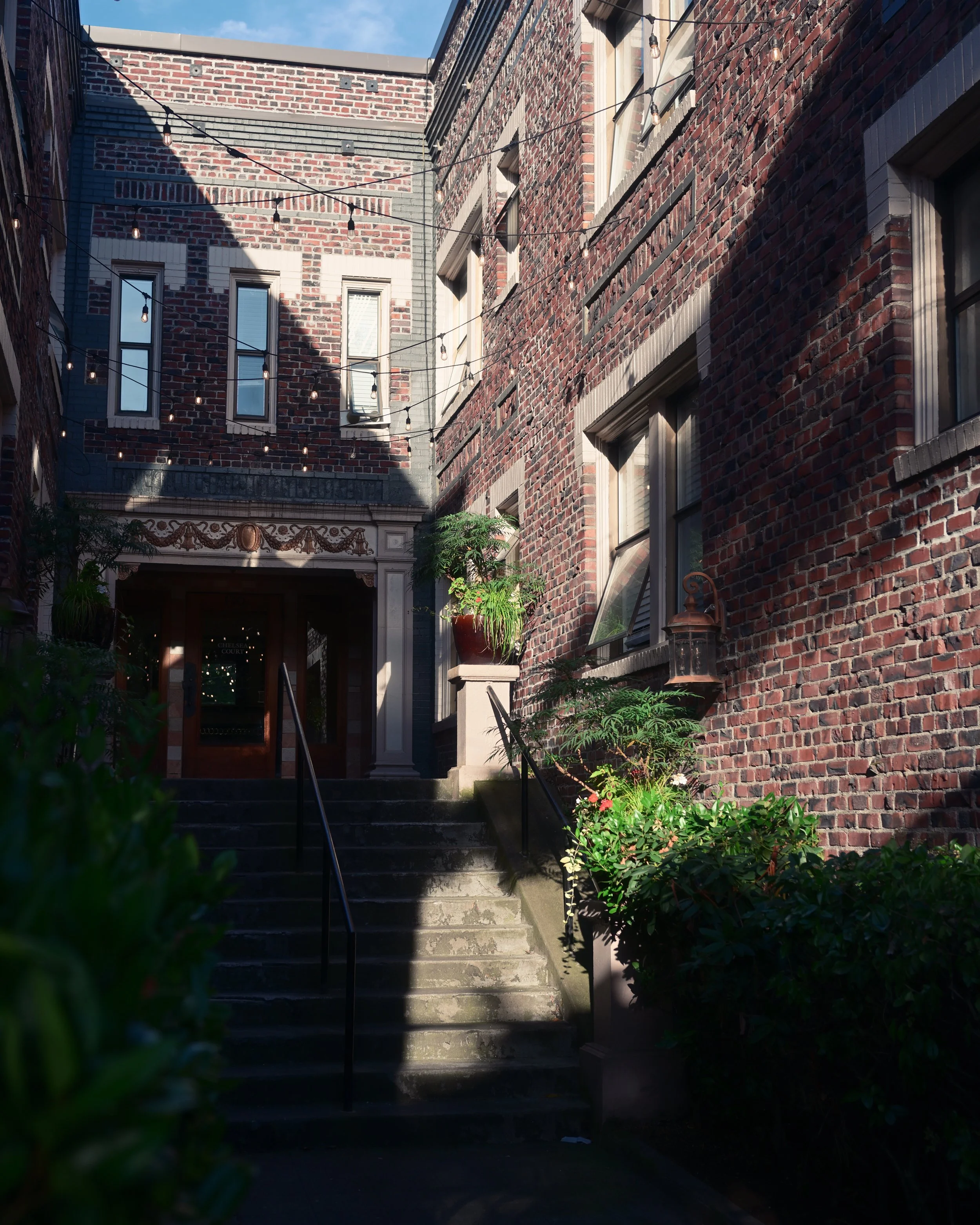 Brick building exterior with stairs leading to a door, potted plants on the sides, hanging string lights above, and windows in the brick wall.