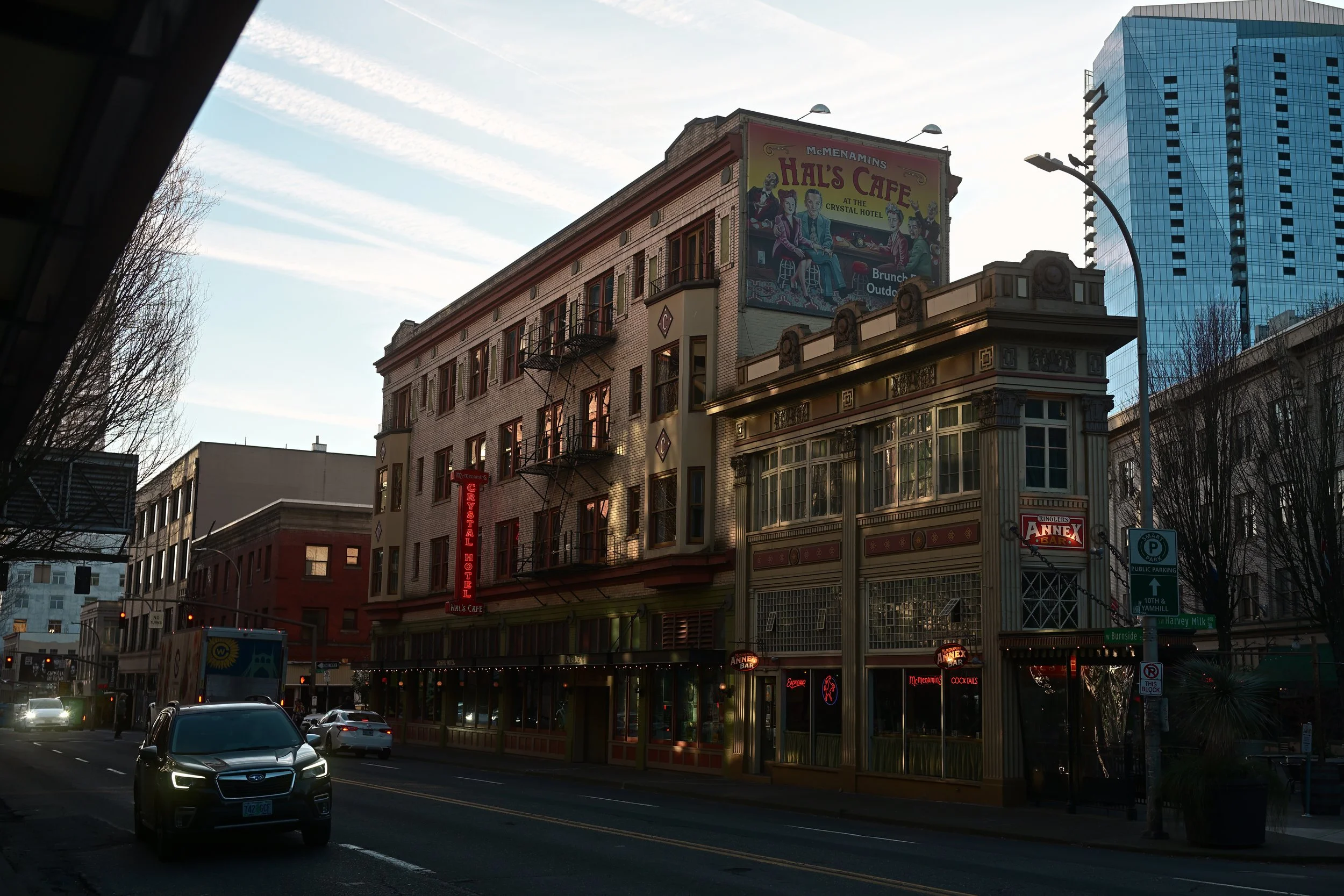 Urban street scene featuring mid-rise historic buildings with signs for Hal's Cafe and other businesses, modern high-rise building in background, cars on the road, and streetlights against a partly cloudy sky.