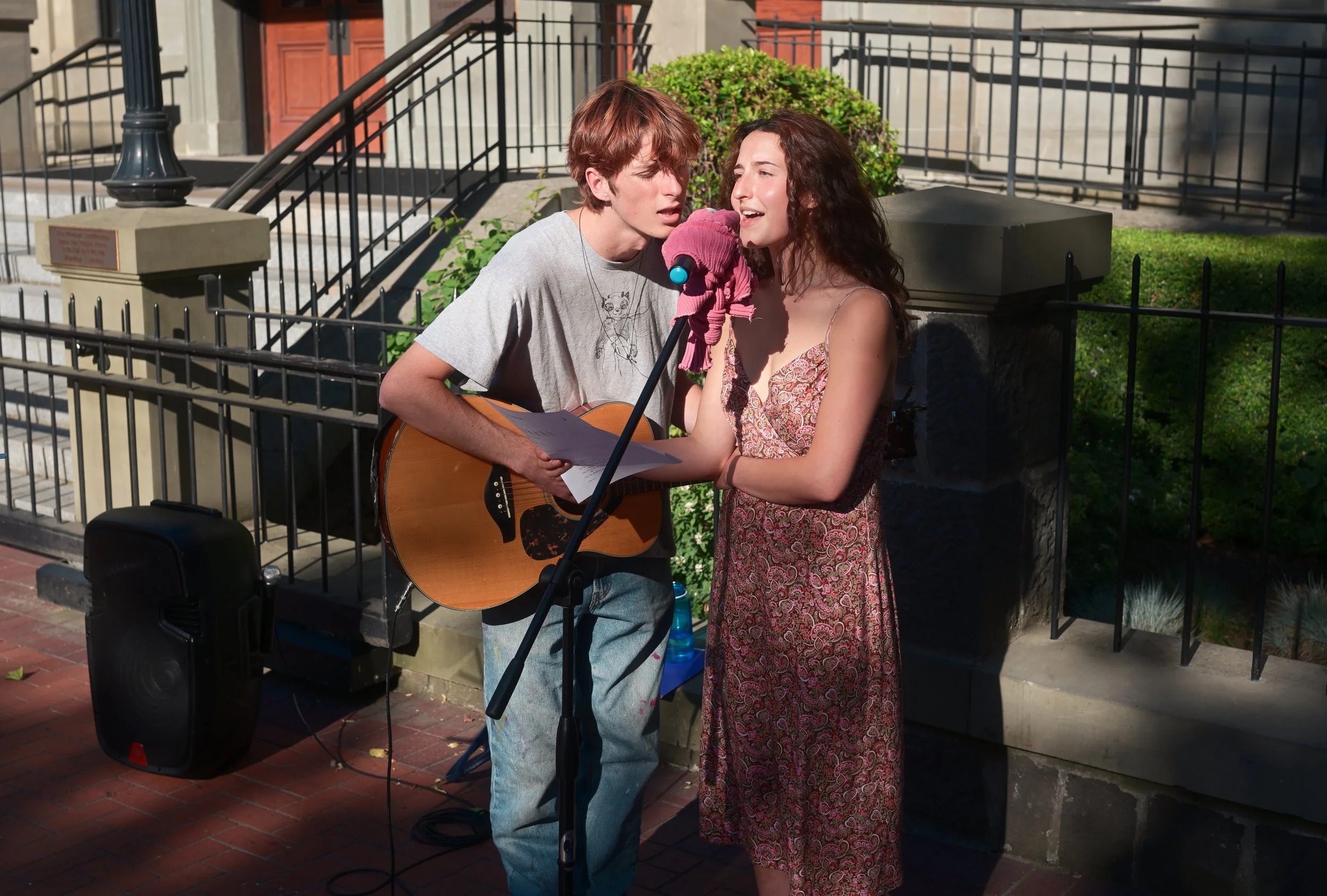 A young man with reddish hair is singing into a microphone while playing an acoustic guitar. A young woman with curly brown hair is standing close to him, singing along. They are outdoors in front of a building with black railings, green bushes, and 