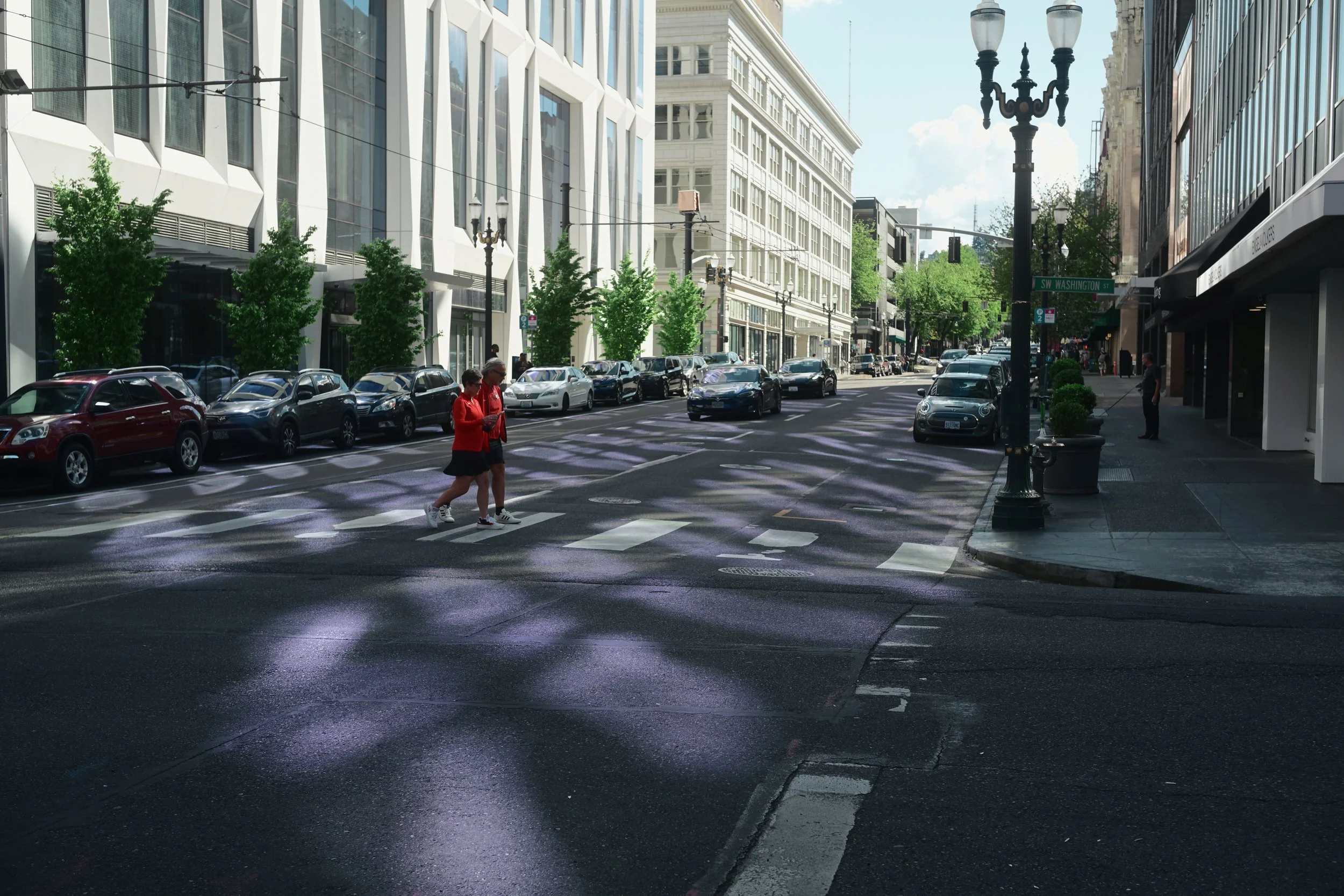 A city street scene showing pedestrians crossing at a crosswalk, parked cars along the curb, modern and historic buildings, street lamps, and a green street sign indicating SW Washington Street.