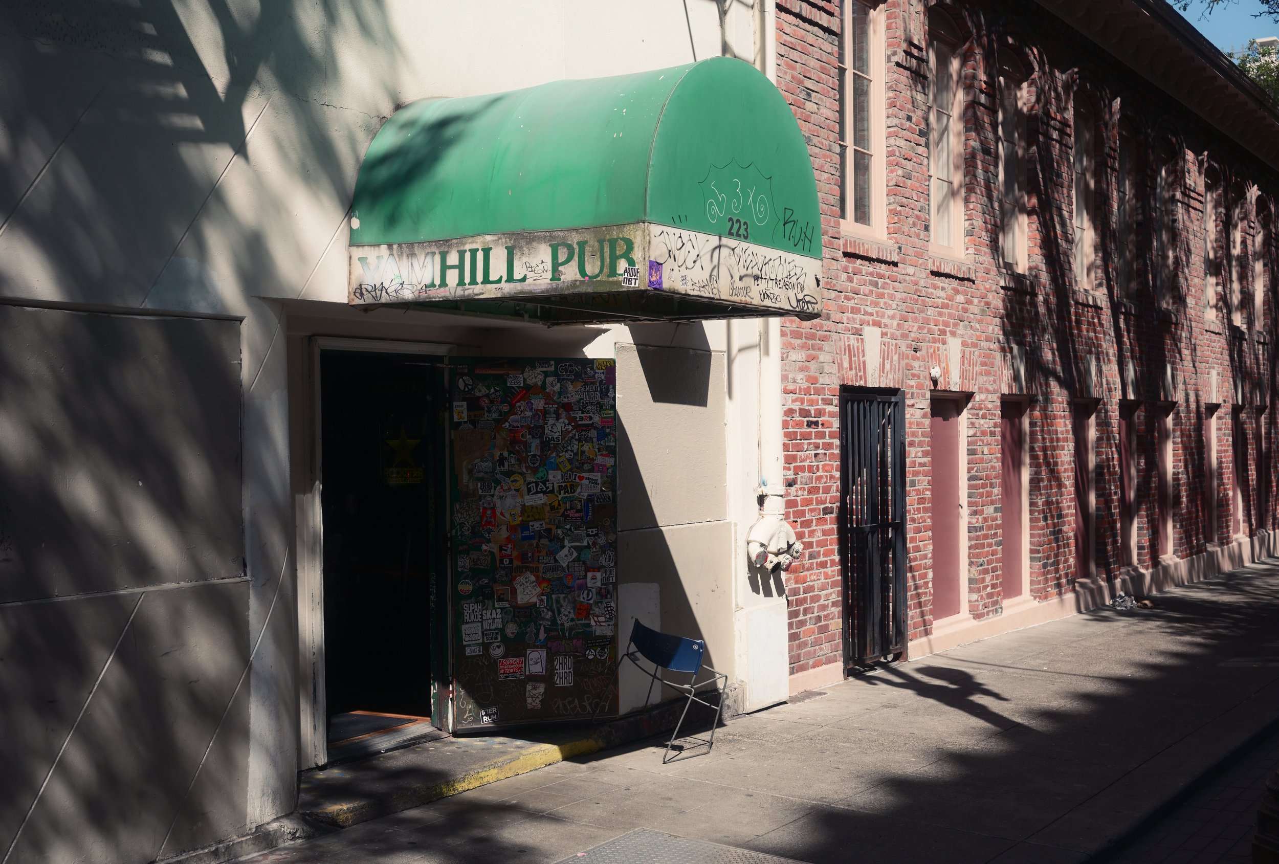 Street scene with a building featuring a green awning over the entrance that has graffiti and stickers. A black chair is placed outside near the door, with shadows from trees cast on the sidewalk and building.