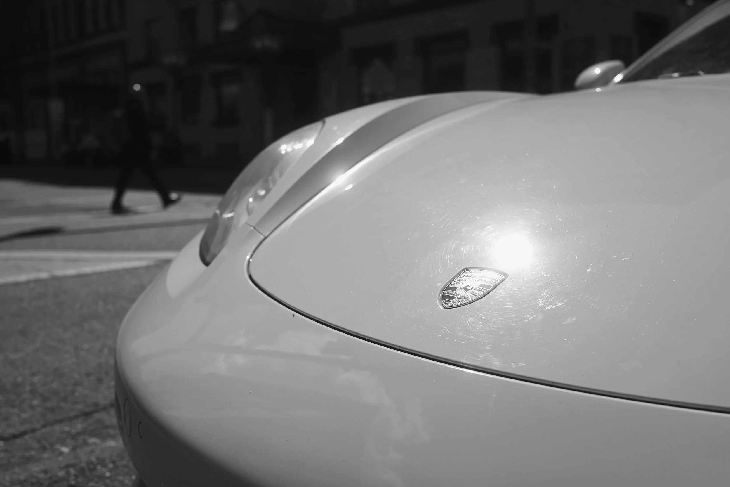 Close-up of the front of a Porsche sports car, showcasing the car's sleek hood and the Porsche emblem. In the background, a person is walking across the street near a row of buildings. The scene is in black and white.