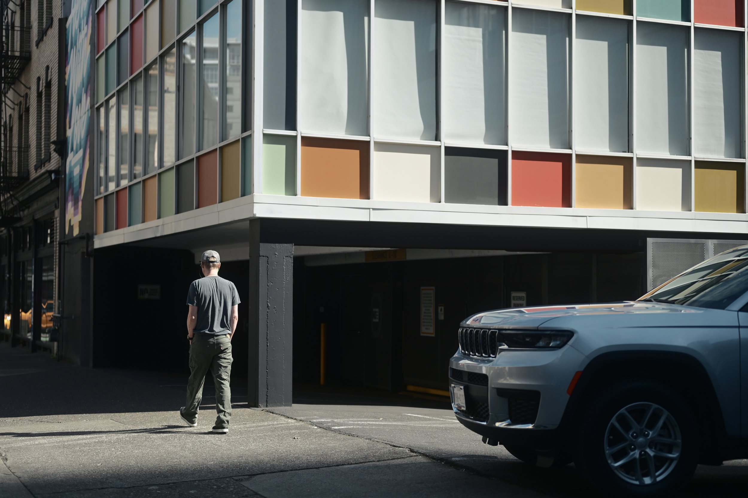 A man with gray hair and wearing a hat walking in a parking lot under a colorful building with multiple windows and a gray car parked nearby.
