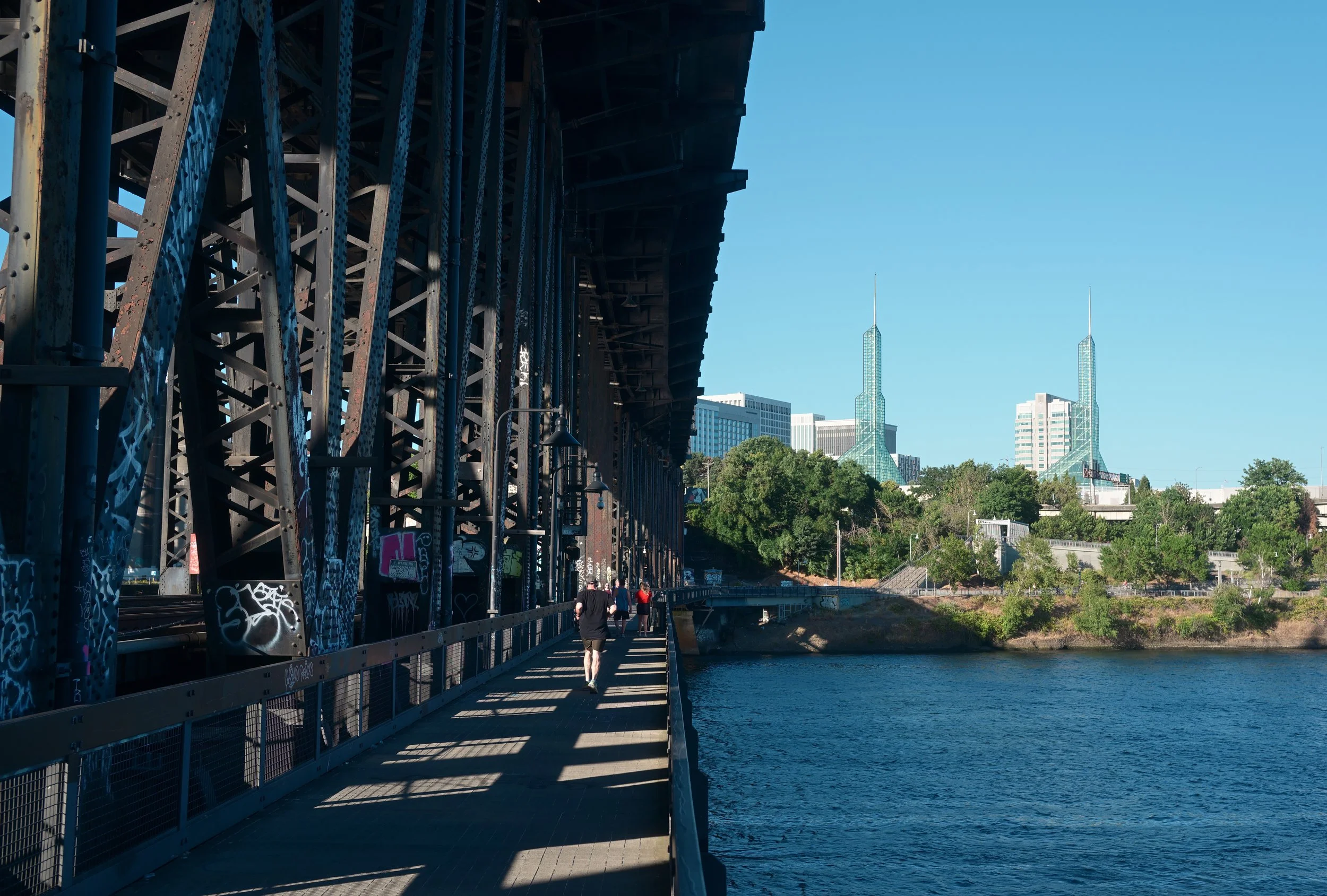 People walking on a bridge over water with city buildings and skyscrapers in the background on a clear, sunny day.