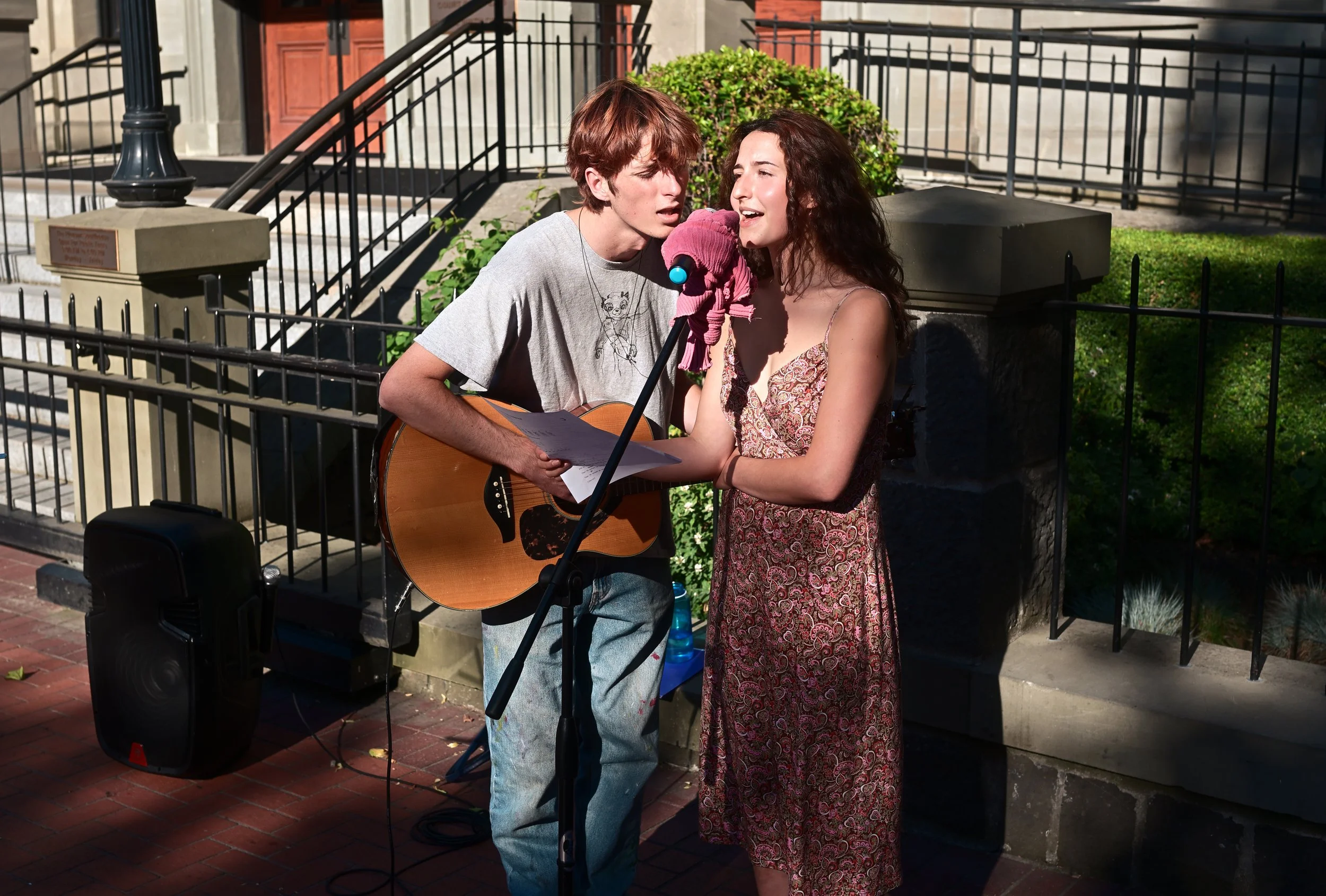 A young man with red hair and a young woman with curly brown hair singing into a microphone while playing guitar outdoors, with a black fence and green bushes behind them.