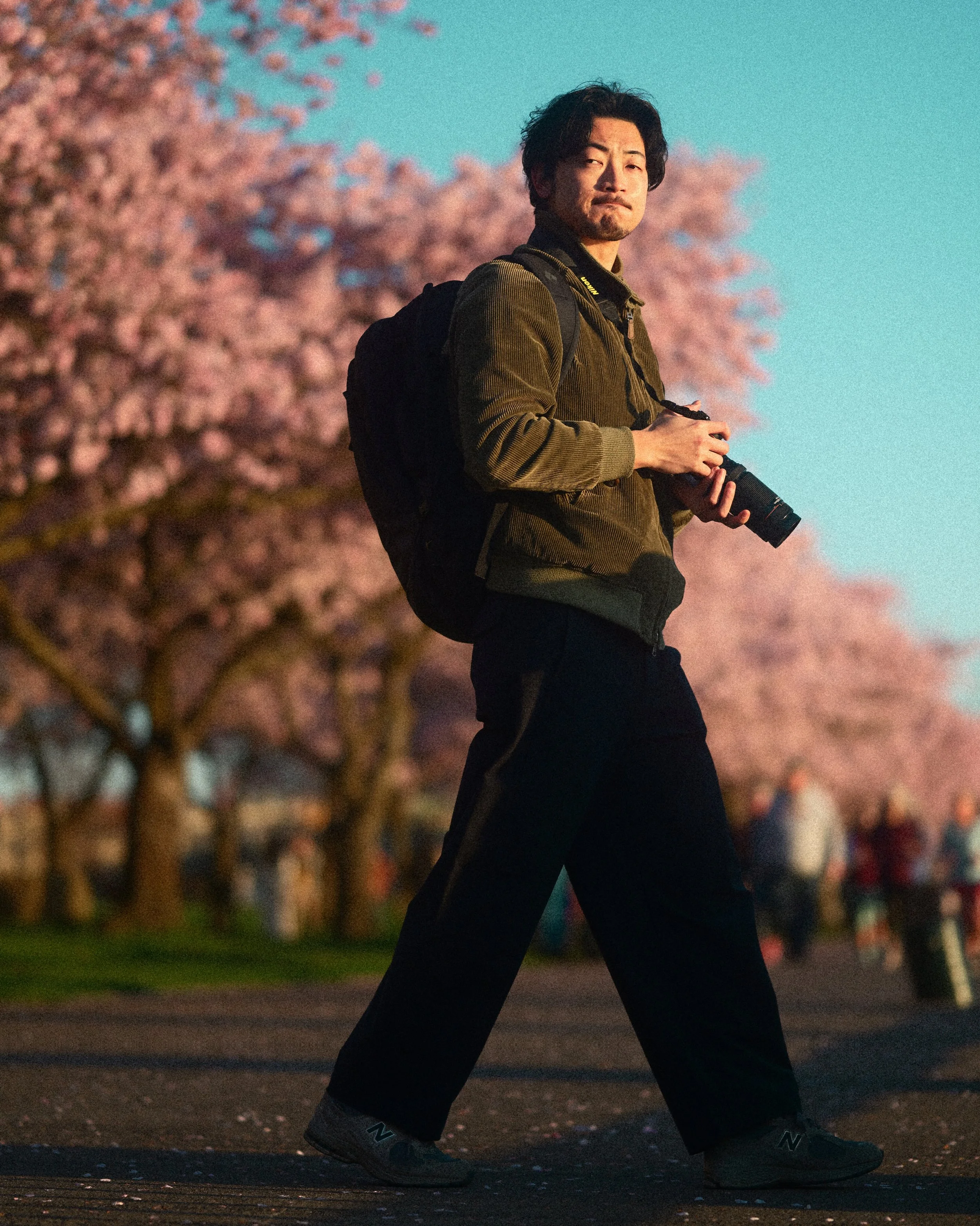 Man wearing a backpack and holding a camera outdoors against a turquoise sky and pink flowering trees