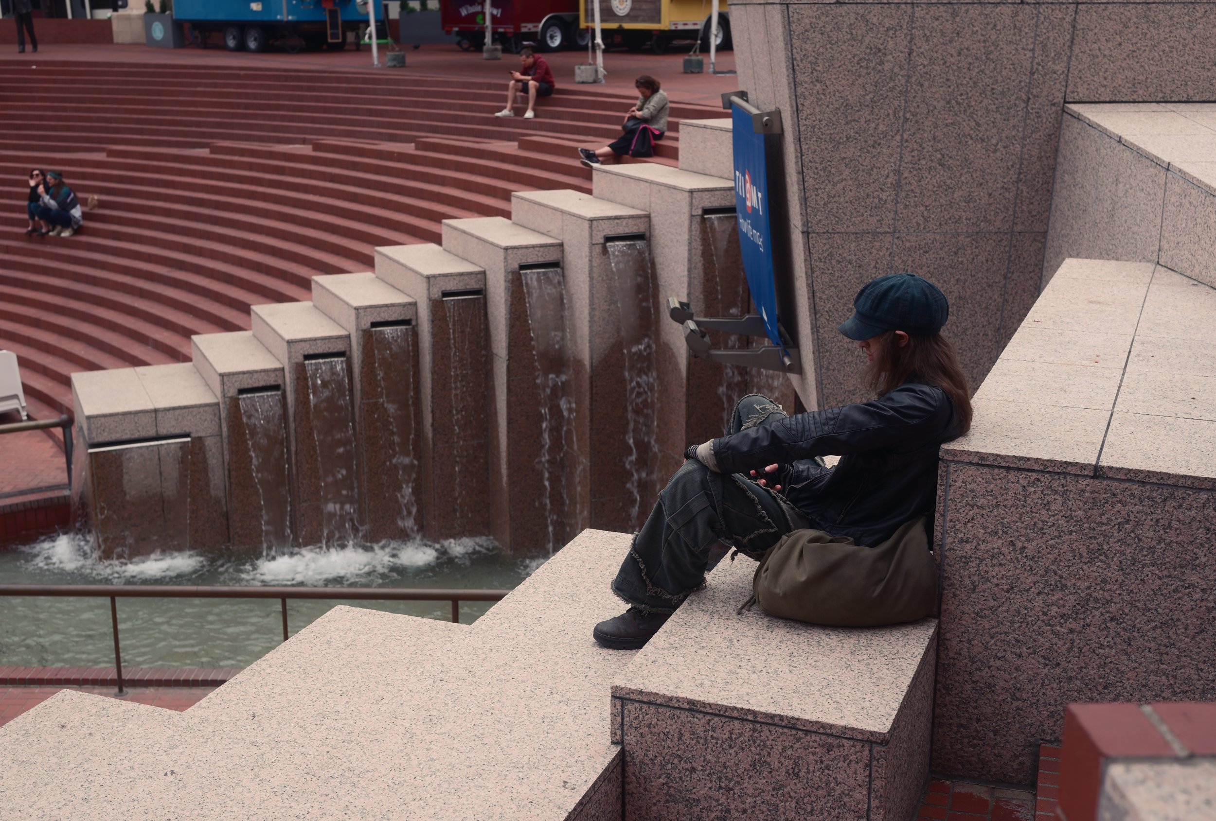 A woman sitting on a stone ledge beside a stepped fountain, looking at her phone, with other people seated on the steps in the background.