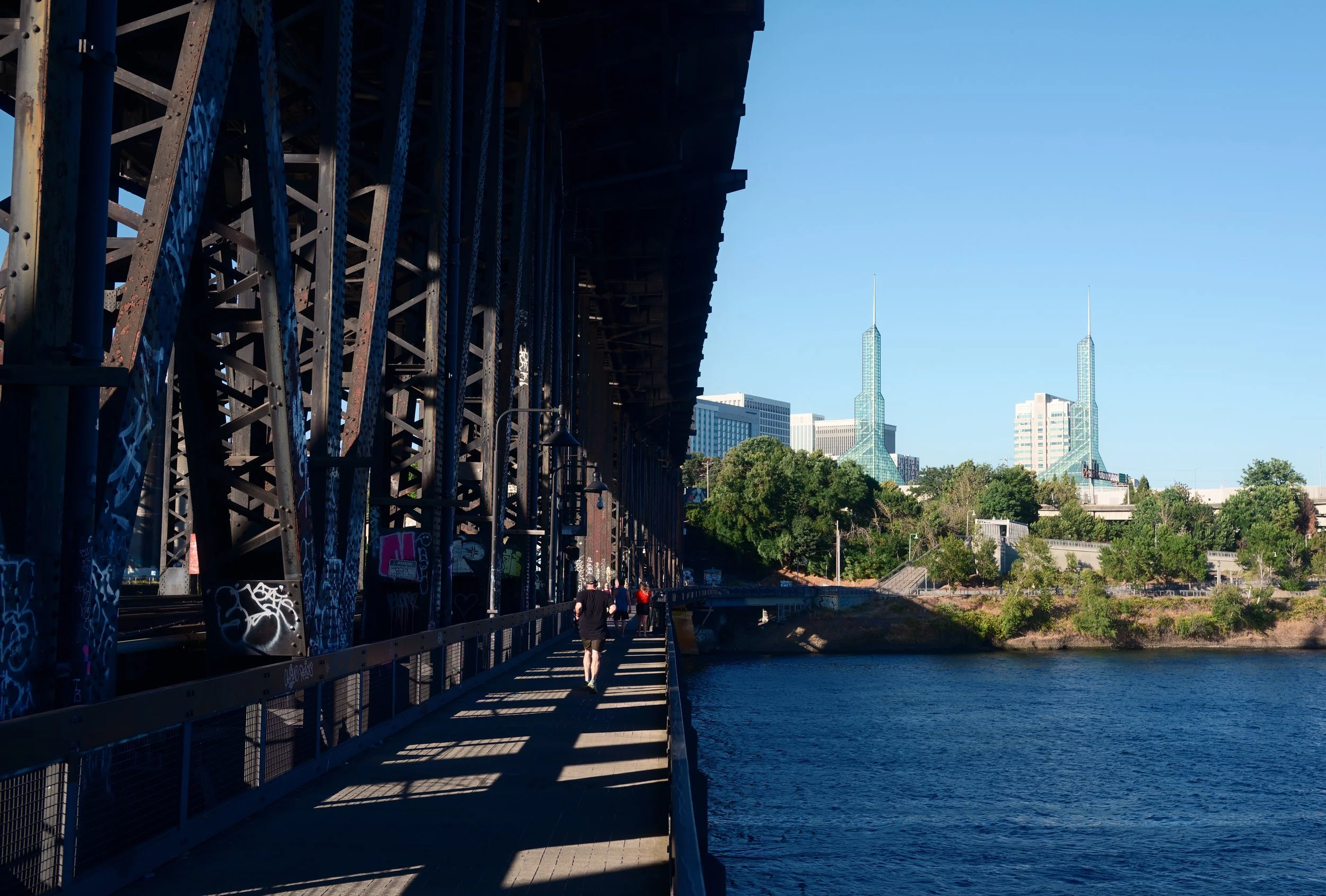 People walking on a riverside under a bridge with metal support beams, graffiti, nearby water, and city skyline with two glass skyscrapers in the background.