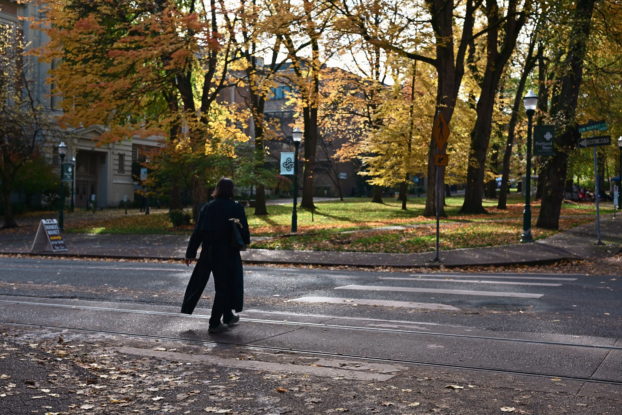 A woman with shoulder-length hair walking across a street at dusk in a park-like area with trees displaying fall foliage, street lamps, and signs indicating it is Portland State University.
