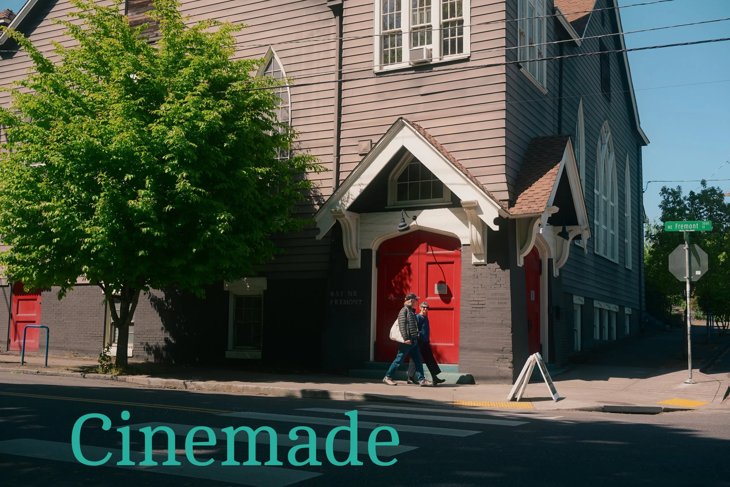 A church building with gray wooden siding, a small gabled entrance with red doors, large windows, and a small cross. Two people are walking past, and there are street signs including one that says NE Fremont.