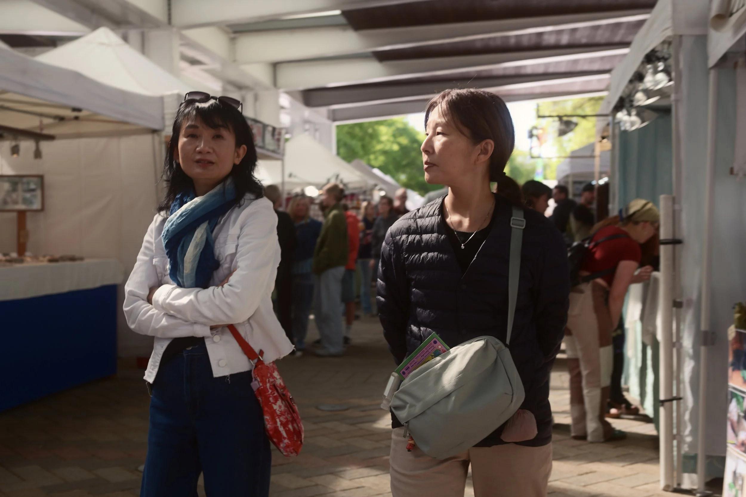 Two women are walking and talking at an outdoor market with white tents and booths. One woman has shoulder-length black hair and is wearing a white jacket and blue scarf. The other woman has short brown hair and is wearing a black jacket. There are other people and green trees in the background.