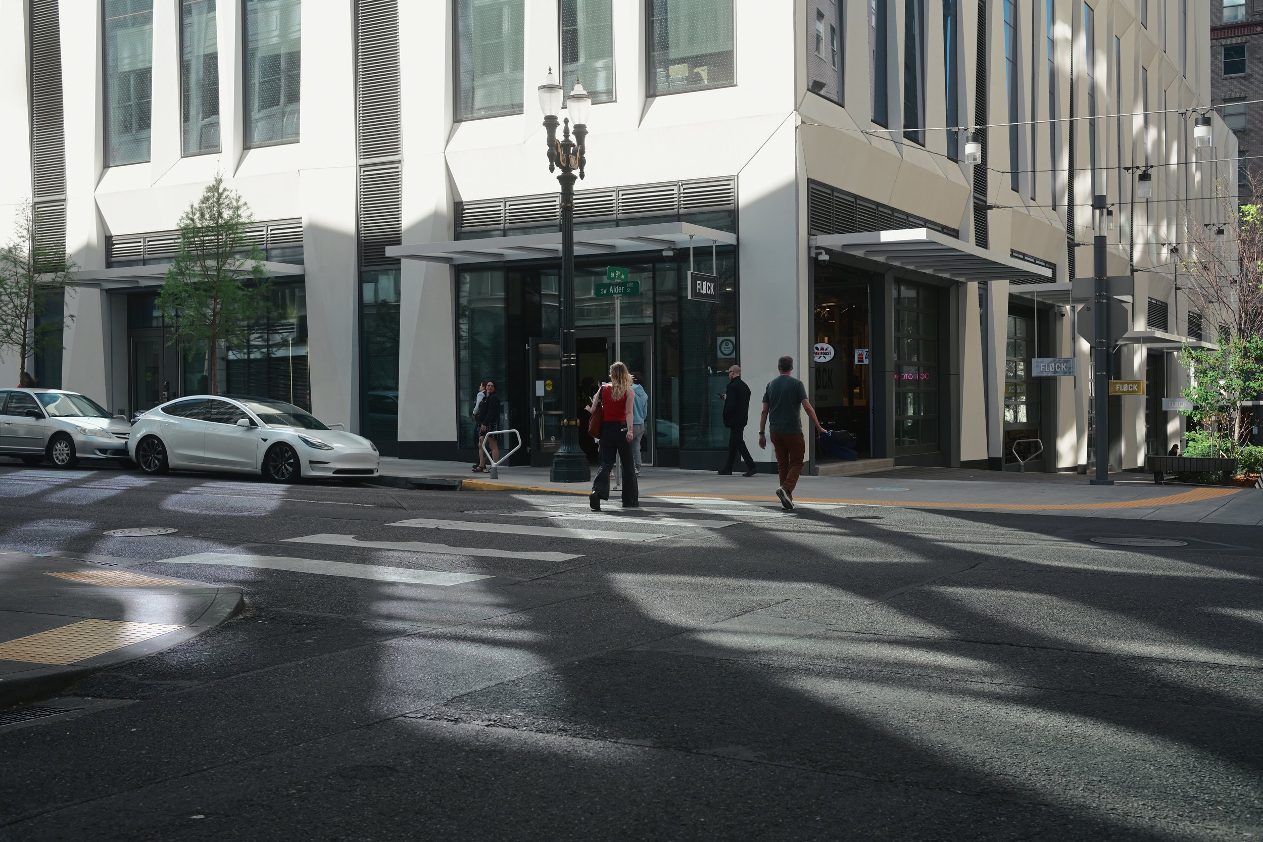 People crossing a street in front of a modern building with large windows and decorative angular panels, cars parked along the curb, and trees with spring foliage. Street signs are visible, including one that reads 'SW Alder'.