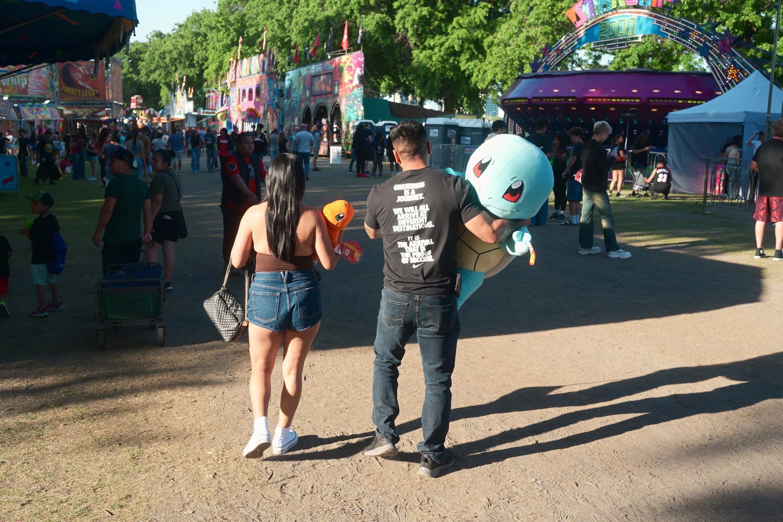 People walking through a colorful carnival with rides, game booths, and food stands under green trees on a sunny day.