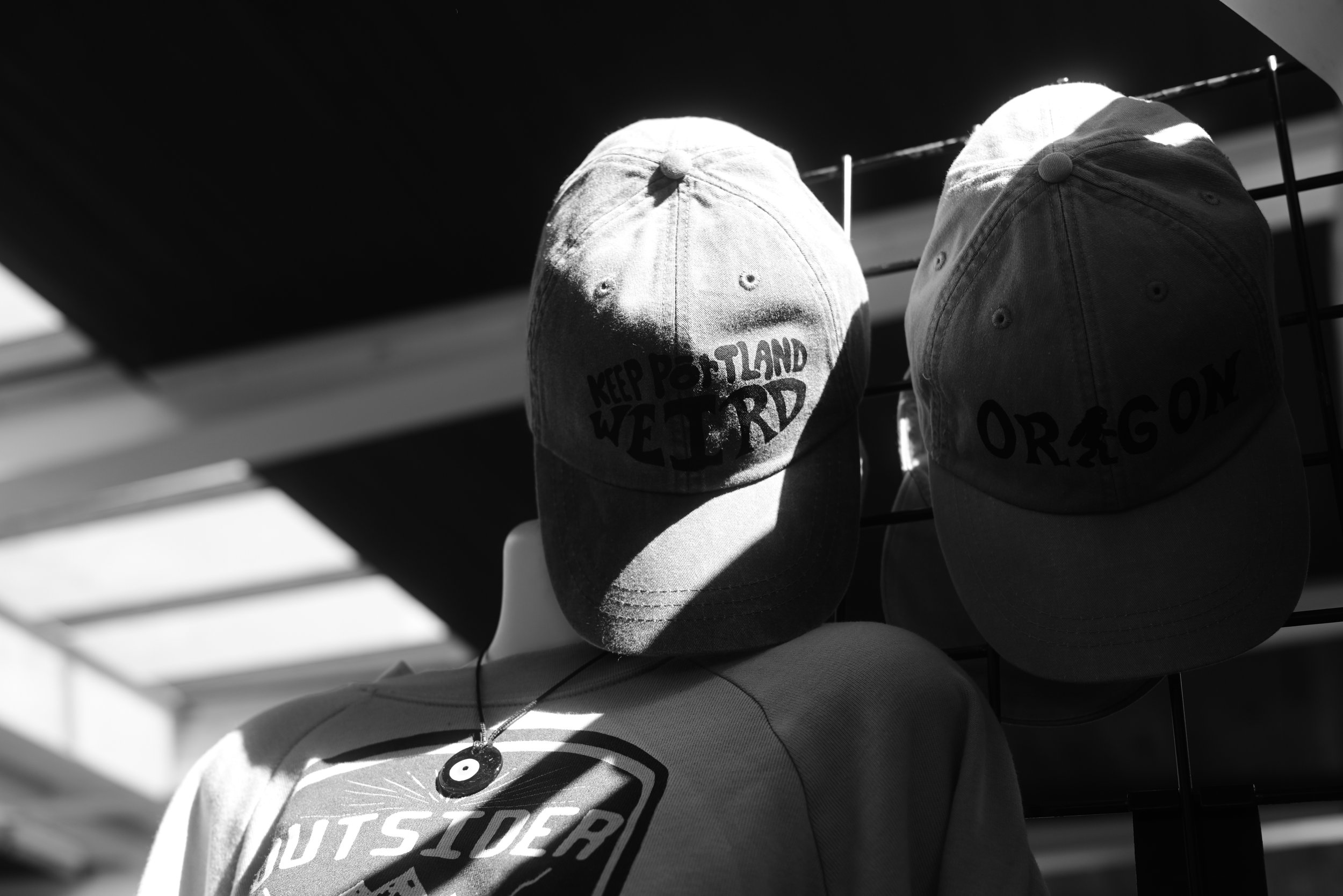 Two baseball caps hanging on a wire rack, with one cap featuring the words 'Keep Portland Weird' and the other with the word 'Oregon.'