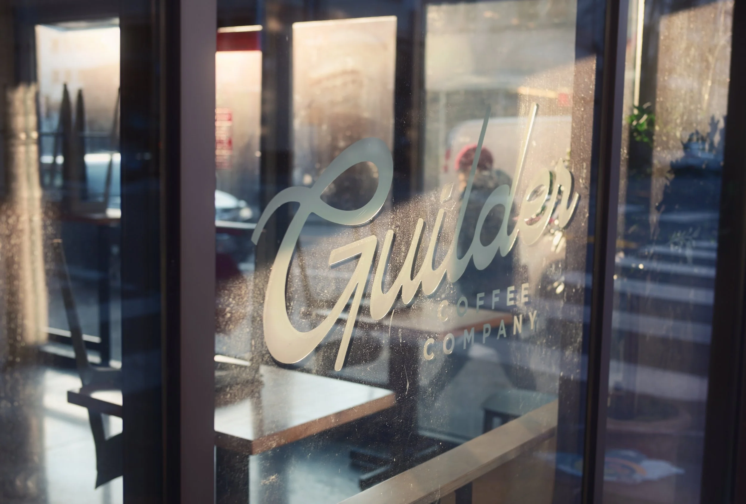 View through a glass window with the logo of 'Gulliver Coffee Company' in white script, showing the interior of a café with tables, chairs, and sunlight coming in from outside.