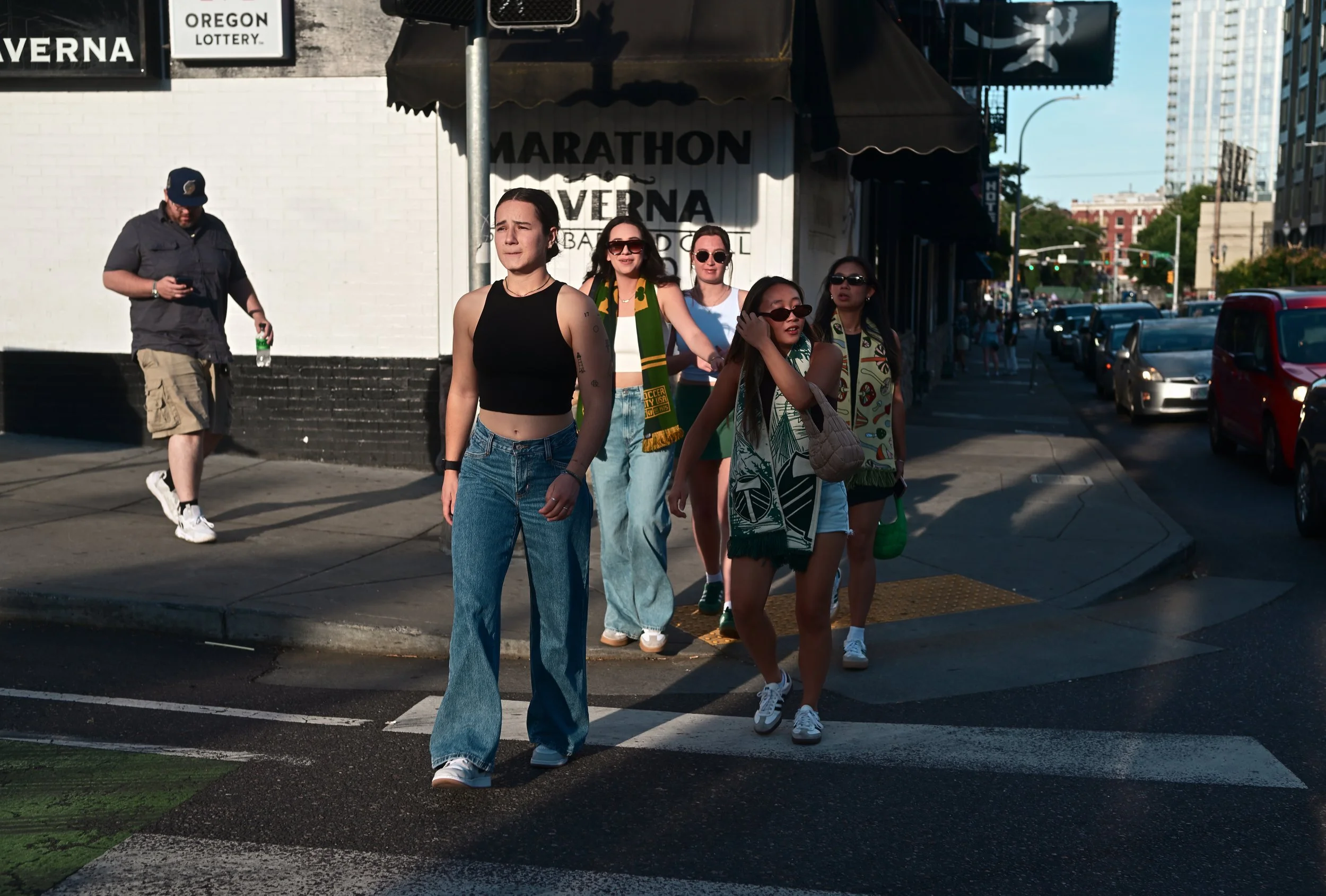 A group of six diverse young women walking across a city street crosswalk on a sunny day, with cars and buildings in the background.