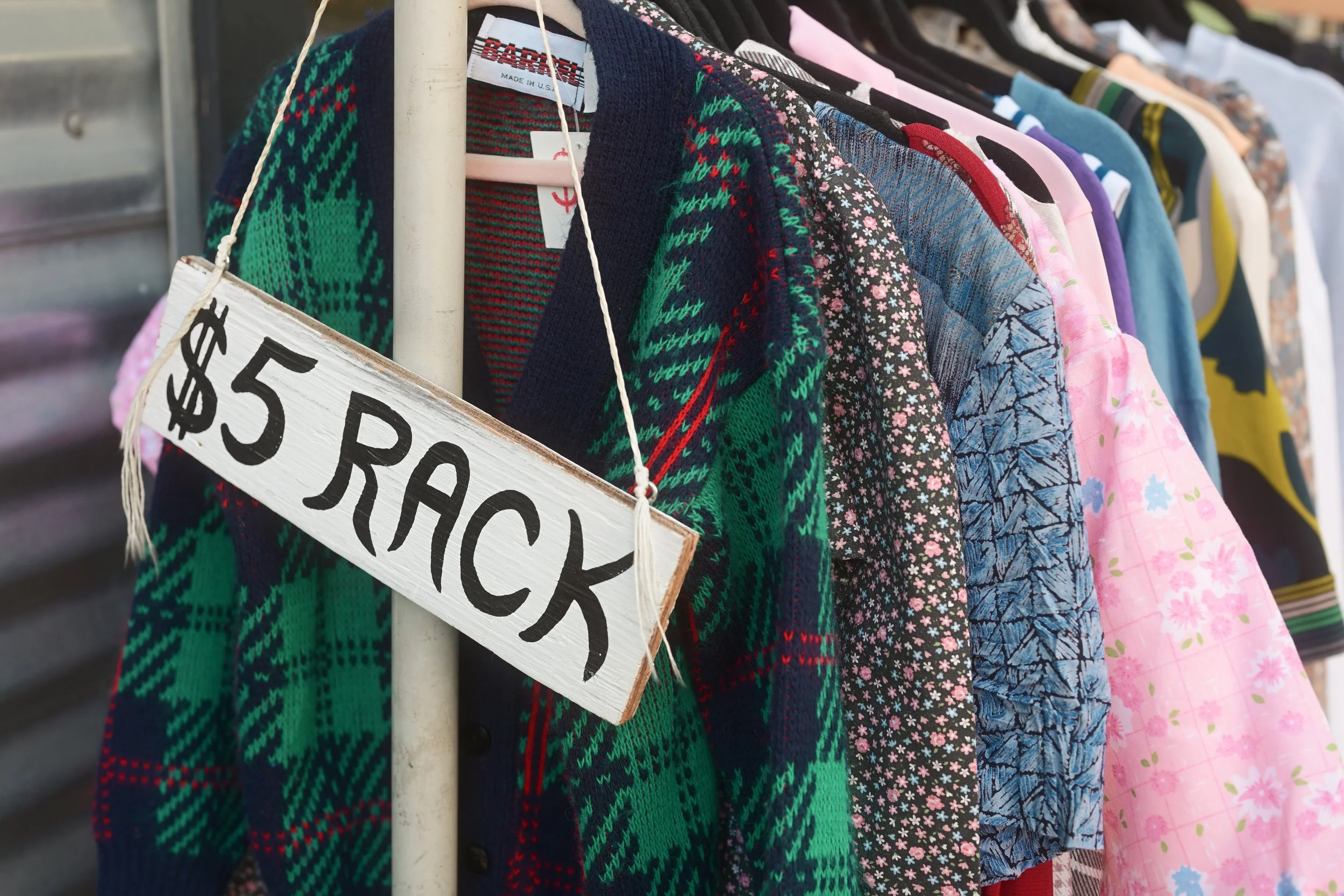 Colorful sweaters and shirts hanging on a clothing rack, with a handwritten sign that says '$5 each' attached to it.