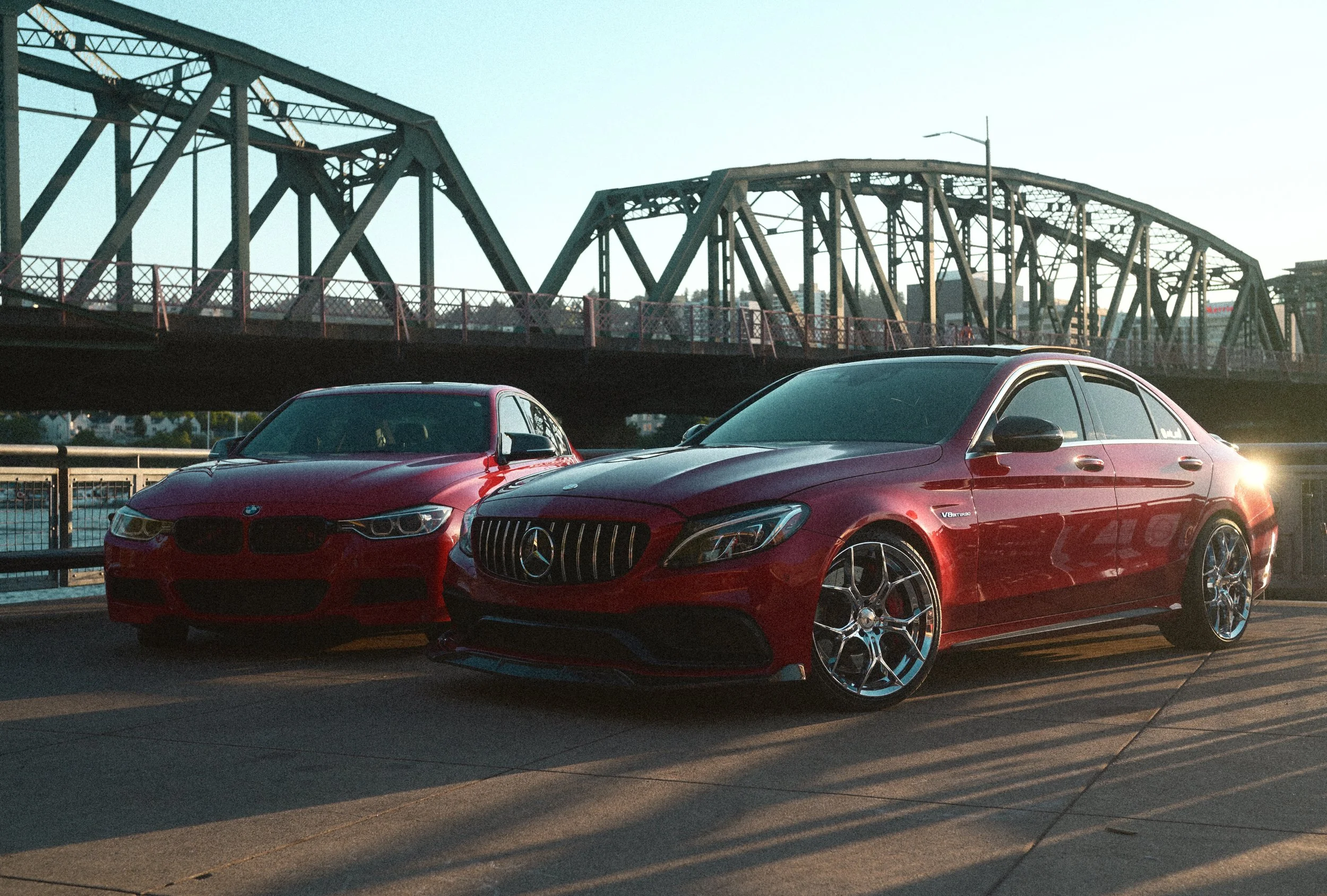Red BMW and Mercedes-Benz luxury sedans parked in front of a steel truss bridge at sunset