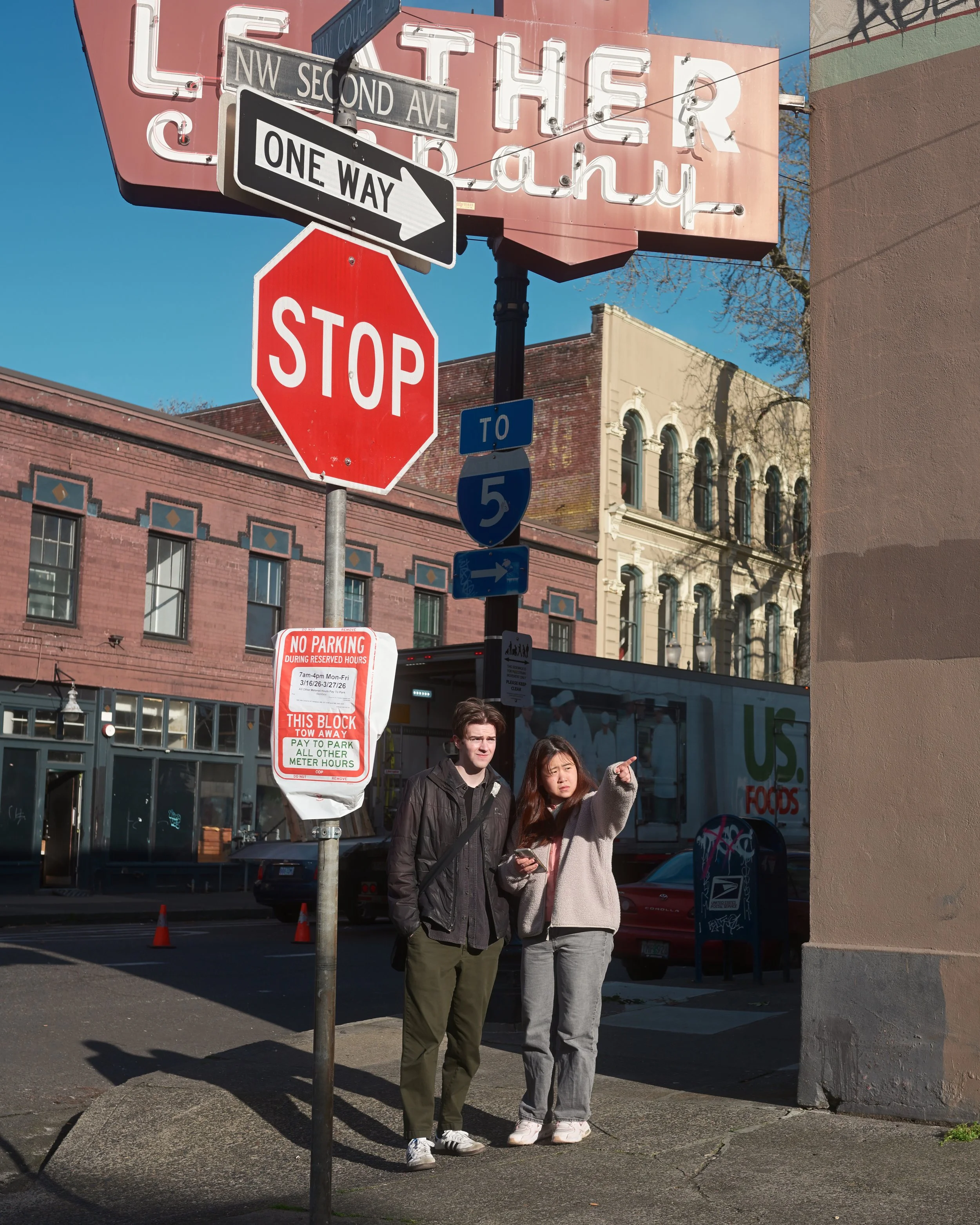 Two young women standing on a city sidewalk near street signs including a stop sign and a one-way sign, with buildings and storefronts in the background during daylight.