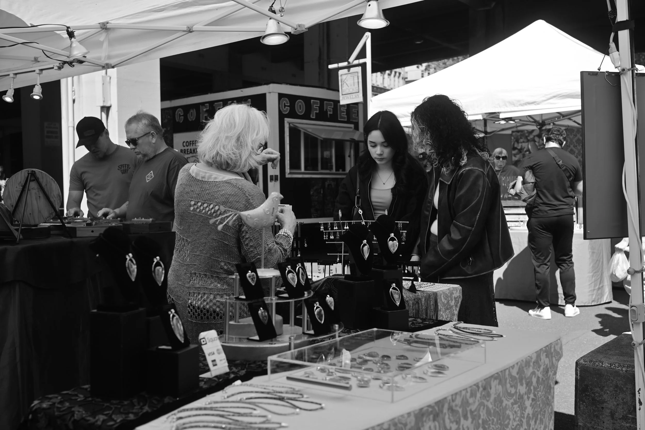 Black and white photo of outdoor market stall selling jewelry and accessories with several people browsing and shopping.