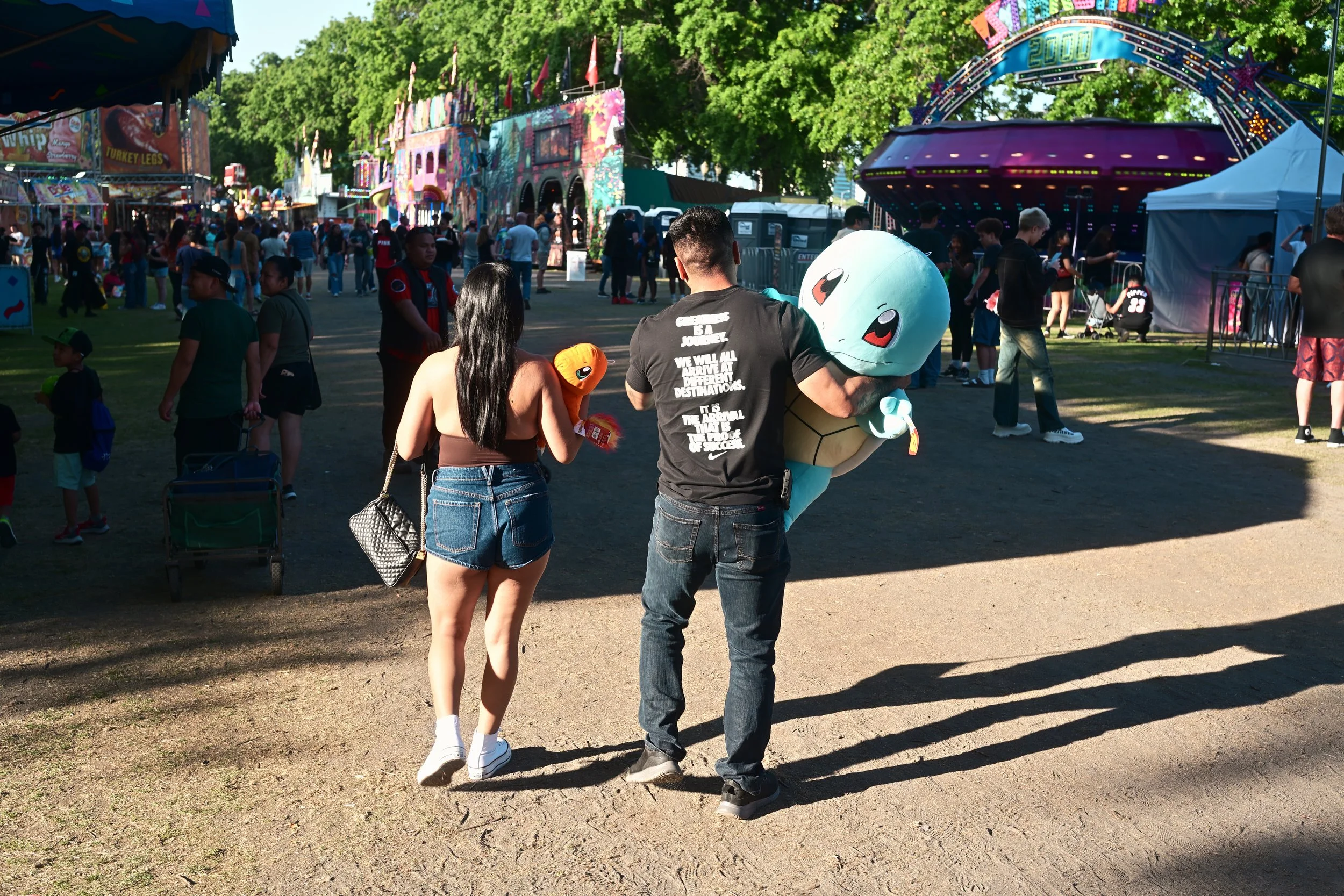 People walking through a lively outdoor carnival or fair with amusement rides, colorful tents, and trees in the background.