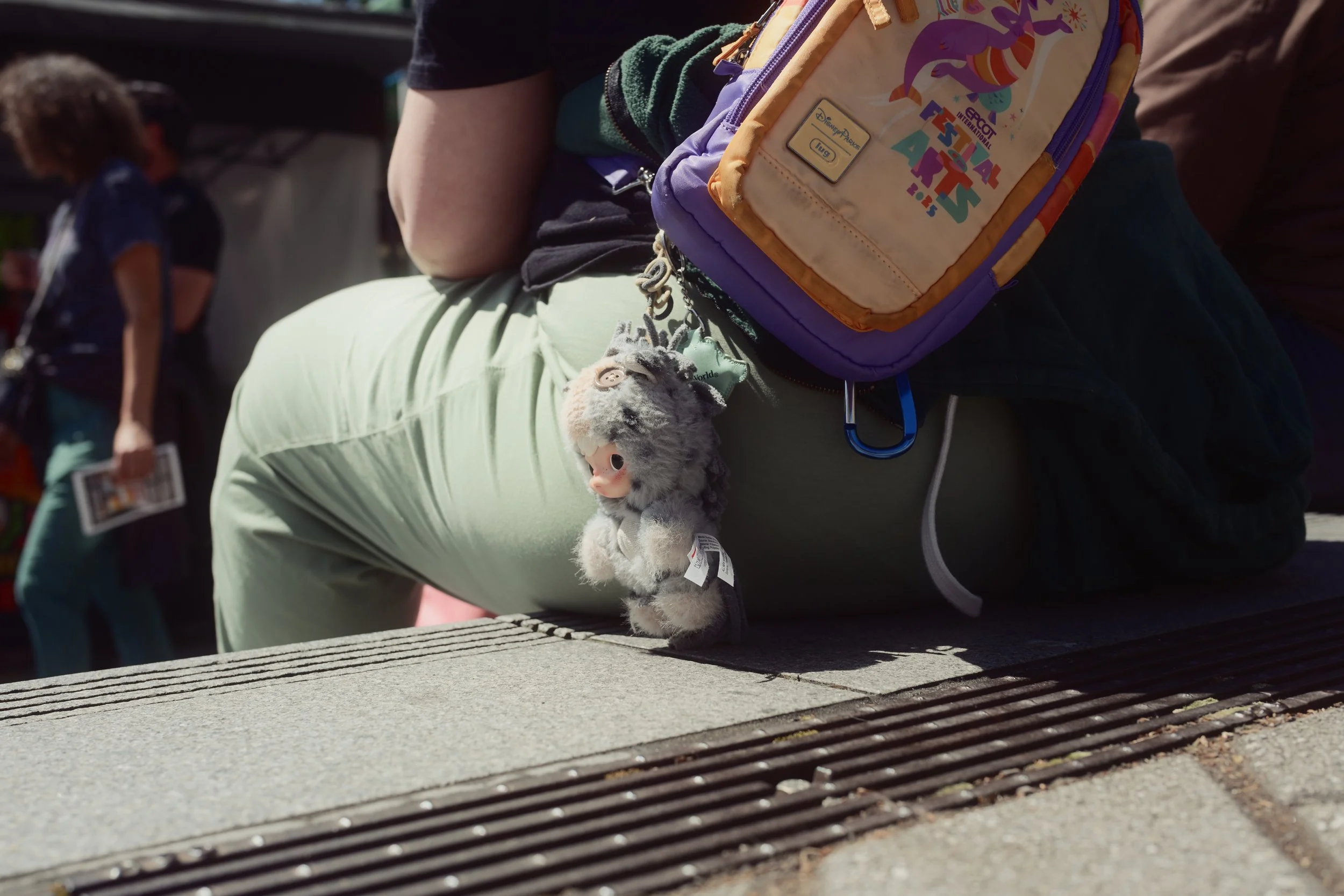 Close-up of a person's lap and backpack with a plush toy attached, seated outdoors on a wooden bench, with other people visible in the background.