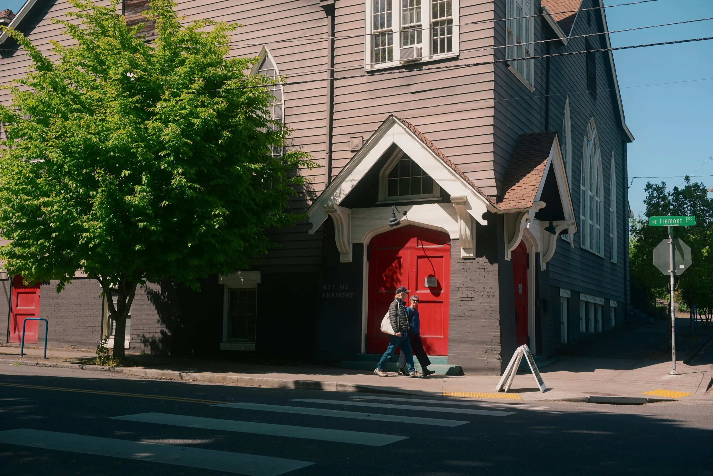 A street corner with a brick church building featuring red doors, white trim, and large arched windows. A green tree partially obscures the building. Two people are walking on the sidewalk. A street sign indicates the intersection with NE Fremont Street.