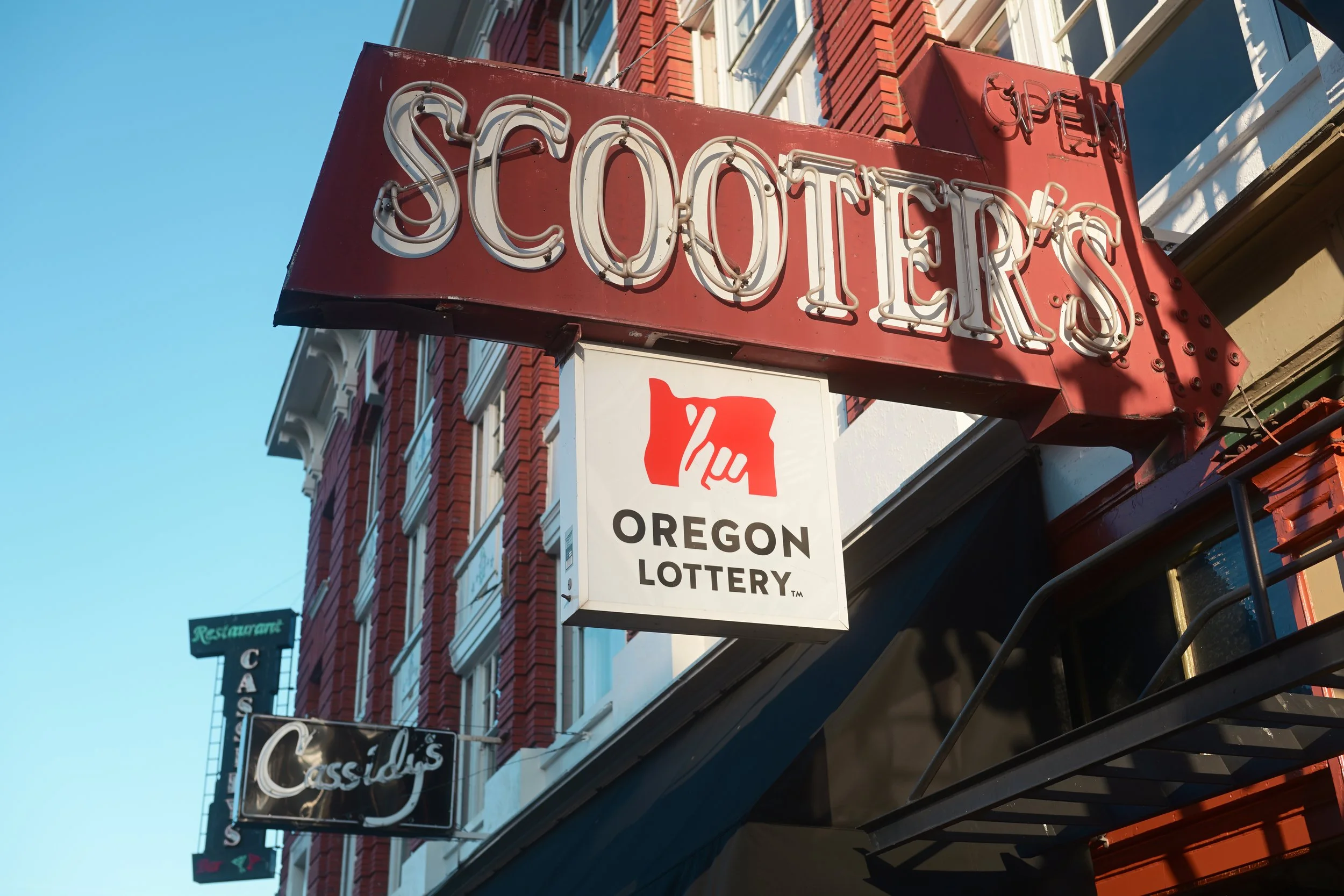 Red neon sign reading 'Shooter's' attached to a brick building, with a white sign below that says 'Oregon Lottery' featuring a red silhouette of the state of Oregon.