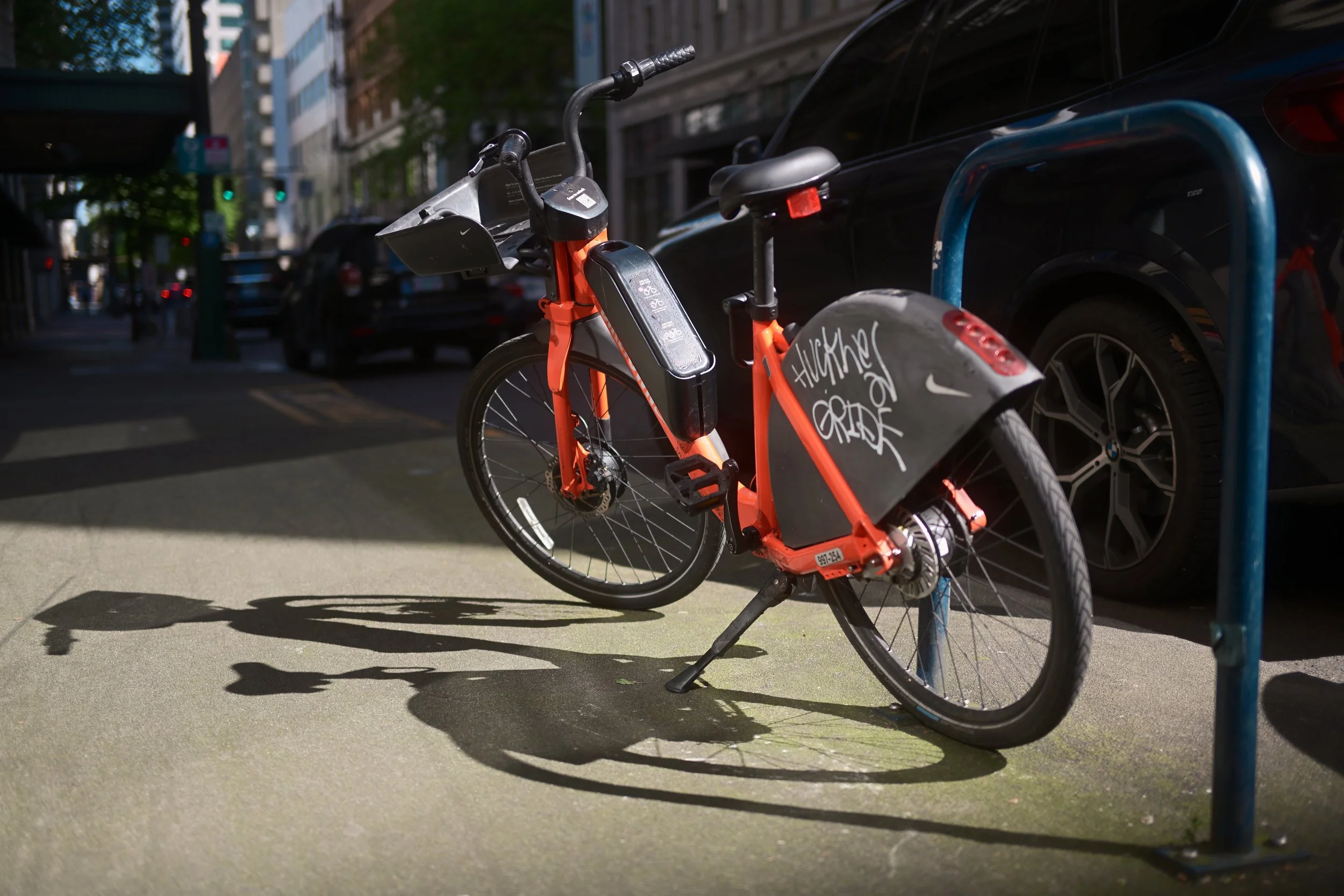 A bright orange shared electric bicycle parked at a bike rack on a city sidewalk, casting shadows on the ground, with cars and buildings in the background.