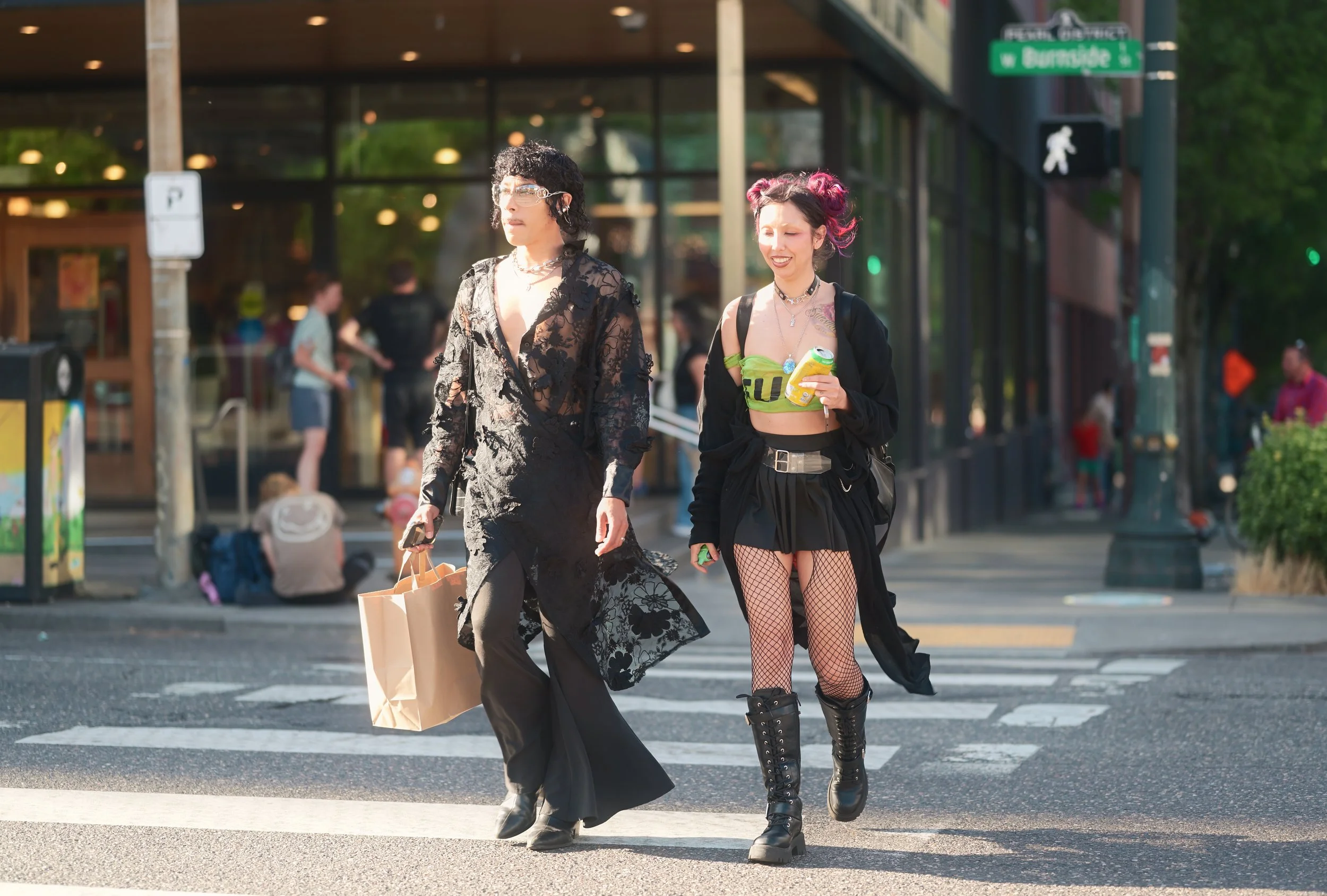 Two young women walking across a crosswalk in an urban area, dressed in alternative fashion with one holding shopping bags, and others in bold, dark clothing, with a backdrop of pedestrians and buildings.