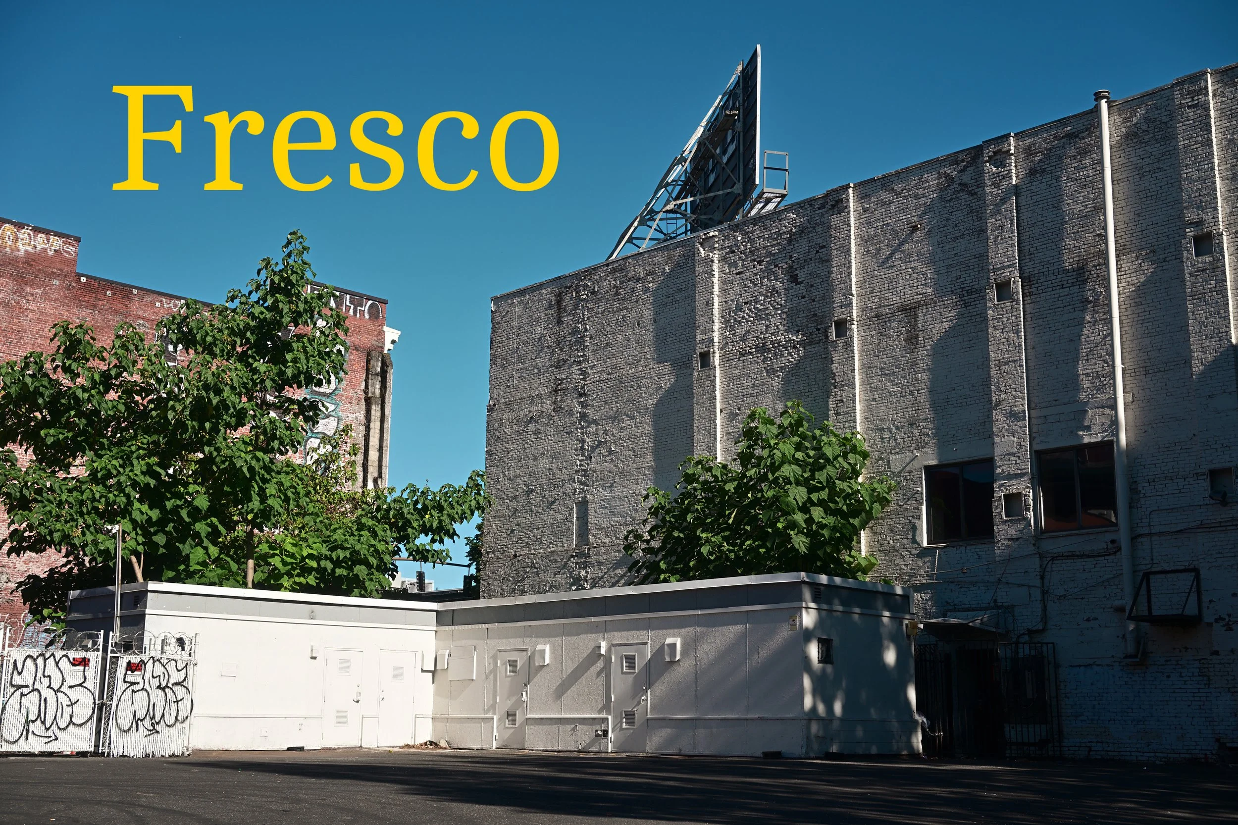 Urban scene with a large brick building, smaller white building, graffiti on the fence, trees, and a blue sky.