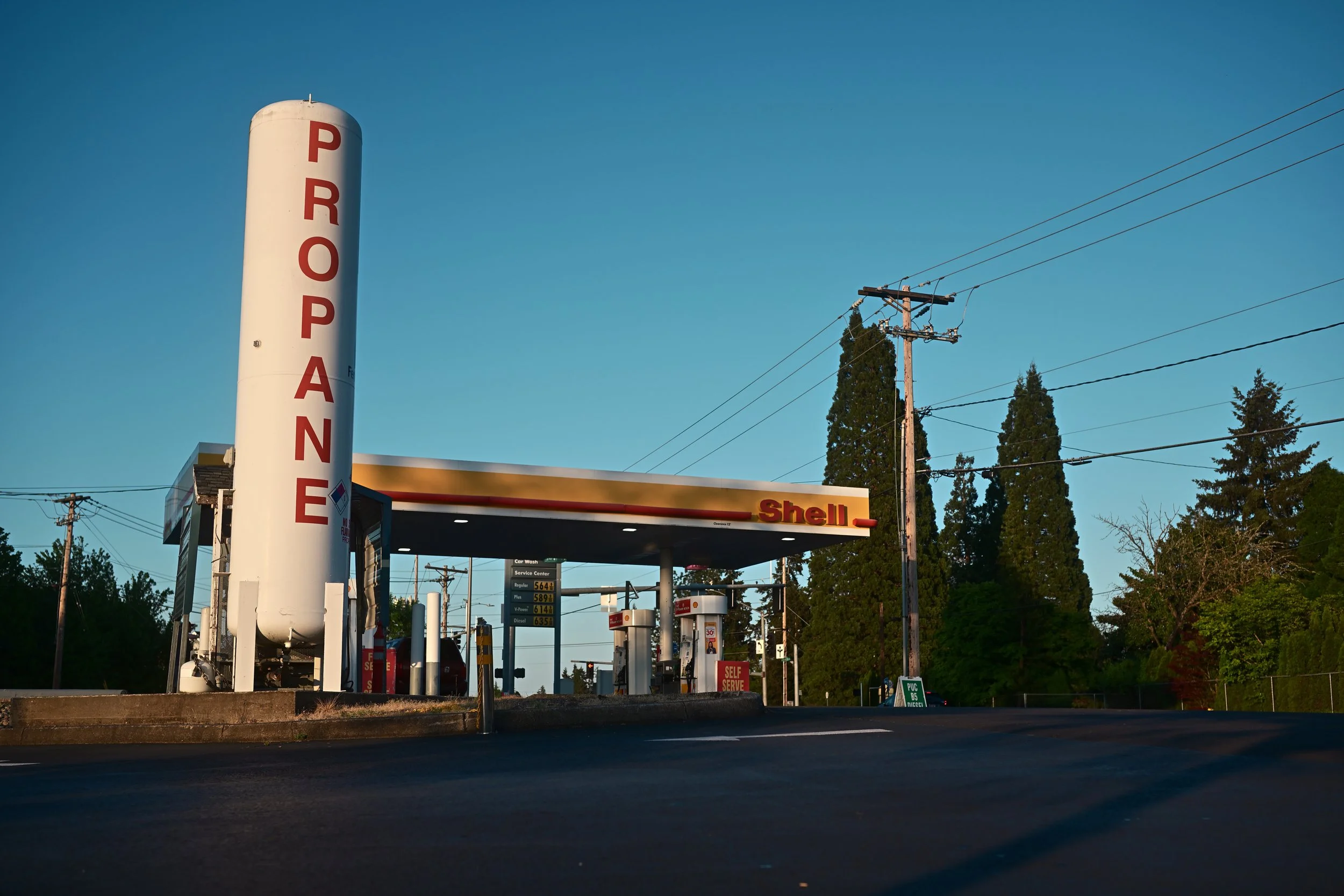 A Shell gas station with a large vertical sign reading 'PROPANE' and gas pumps in the foreground, utility poles with power lines, trees in the background, and a clear blue sky.