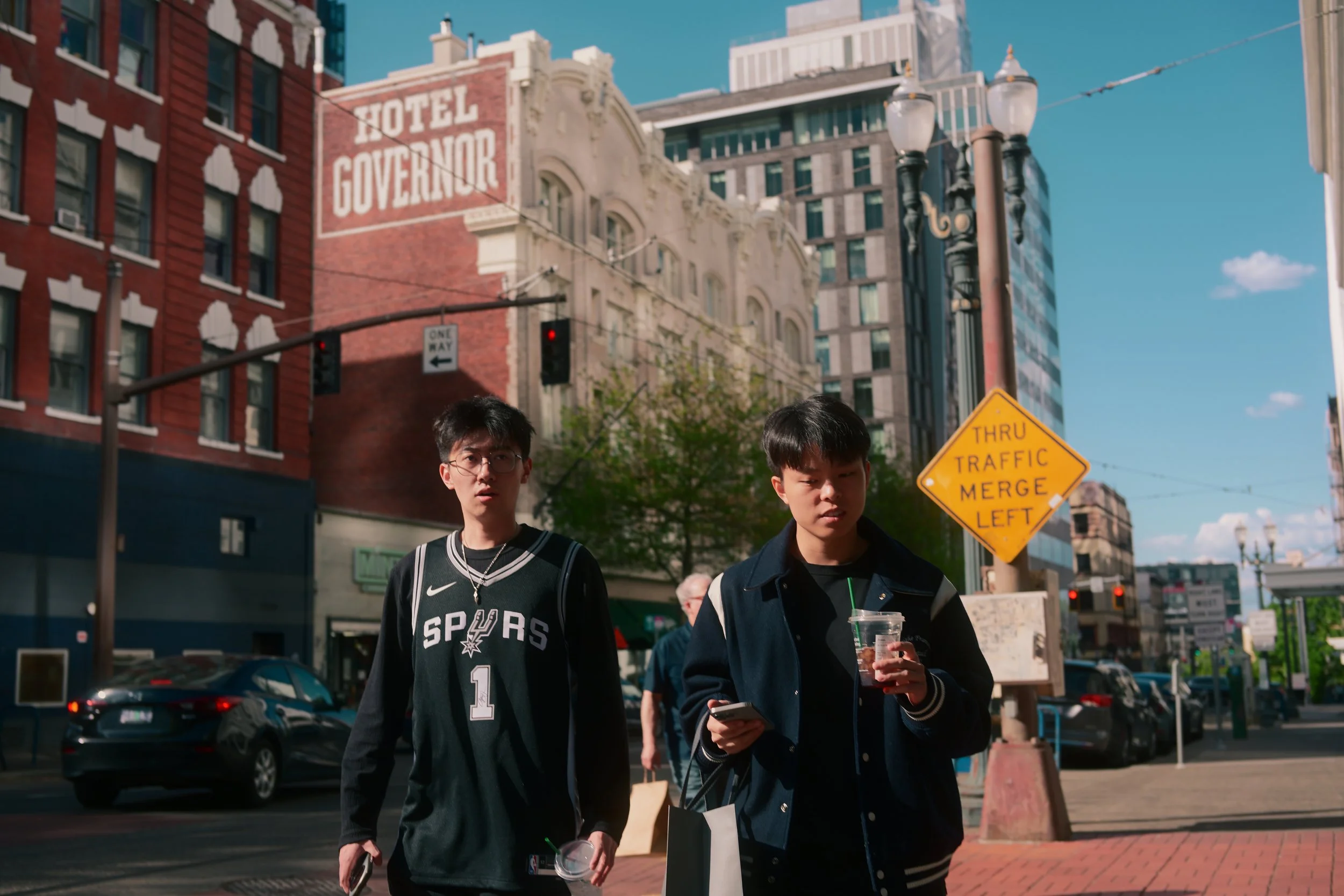 Two young men walking on a city street, one wearing a San Antonio Spurs basketball jersey, the other looking at his phone, with buildings, a hotel sign, traffic lights, and a yellow traffic merge sign in the background.