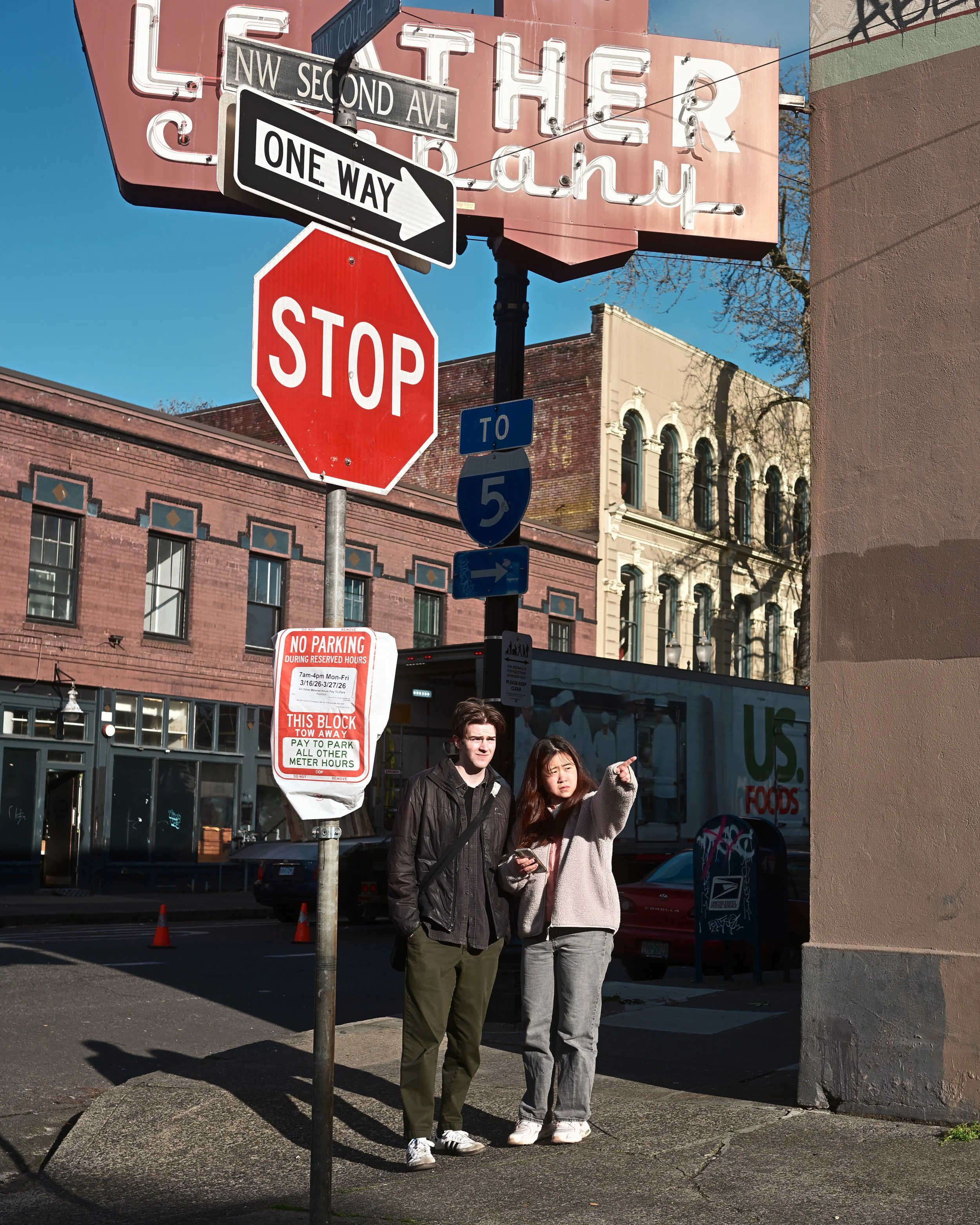 Two young women standing on a city sidewalk, with one of them pointing to something while the other looks on. Behind them are street signs, including a stop sign and directional signs, with buildings and a bus in the background under a clear, sunny s