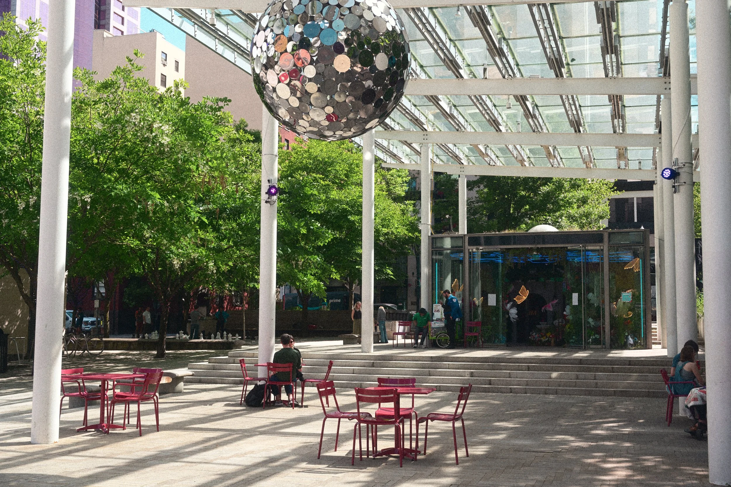 Outdoor urban plaza with red café tables, white columns, glass canopy, trees, and a large mosaic-covered spherical sculpture