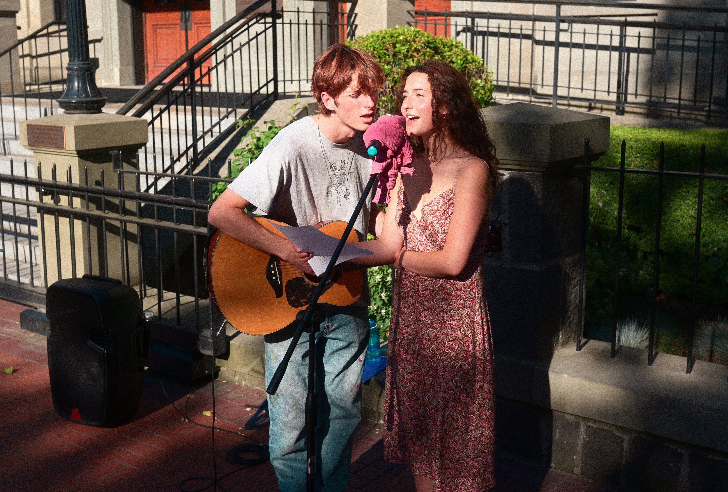 Two street musicians perform outdoors, with a guitarist and singer sharing a microphone beside a black speaker.