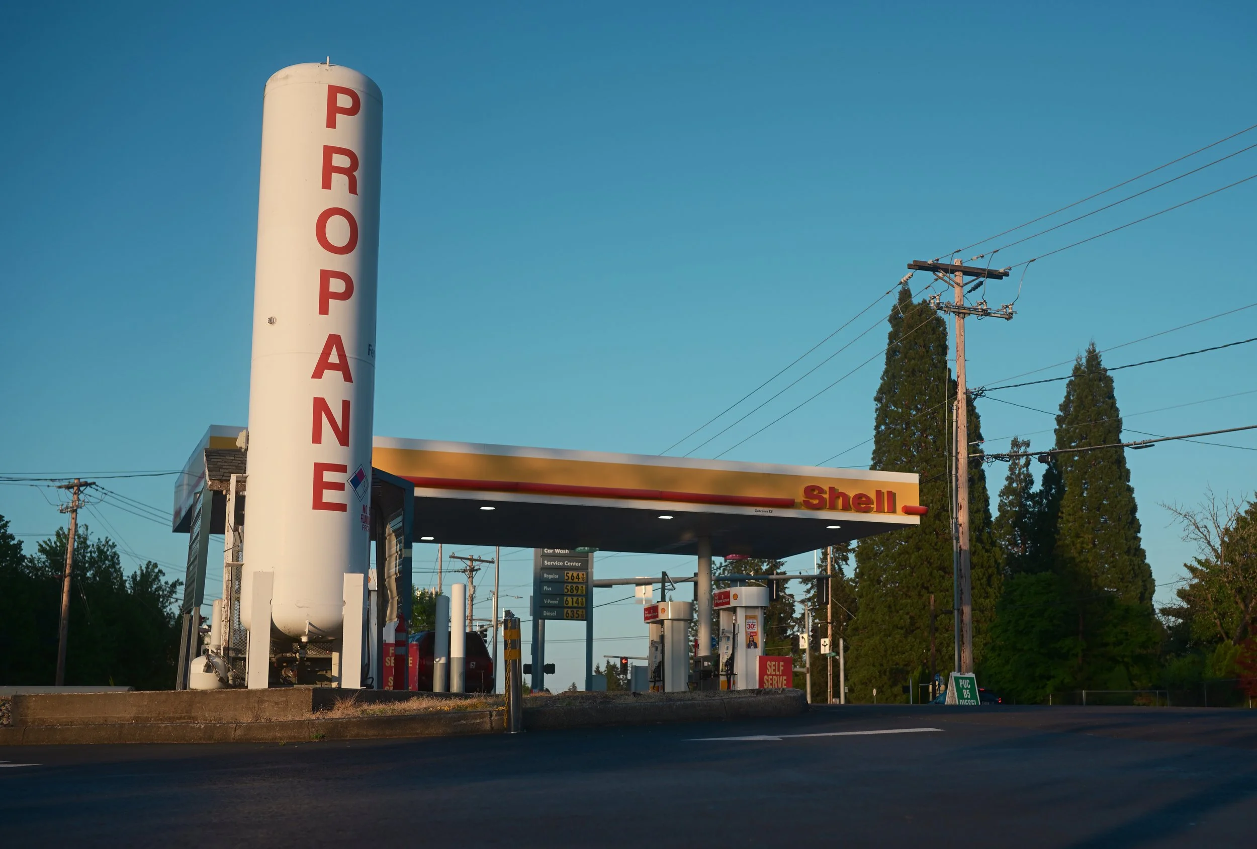 A Shell gas station with fuel prices displayed on a sign, tall white fuel storage tank with red 'PROPANE' letters, and utility poles with power lines against a clear blue sky. Trees are visible in the background.