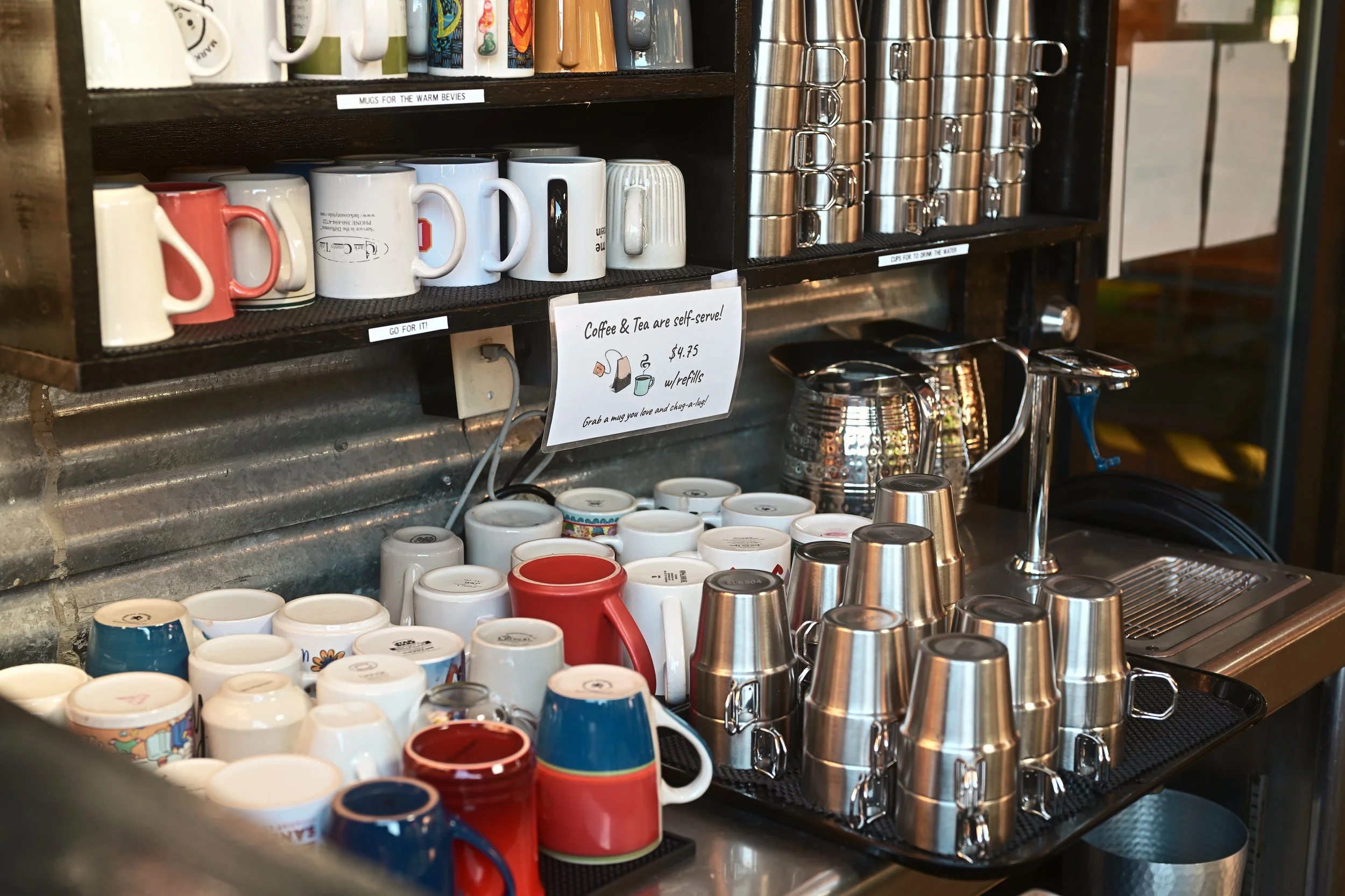 Coffee shop or cafe counter with stacked mugs, cups, and stainless steel containers for drinks. Signs indicating self-serve coffee and tea for $4.75.