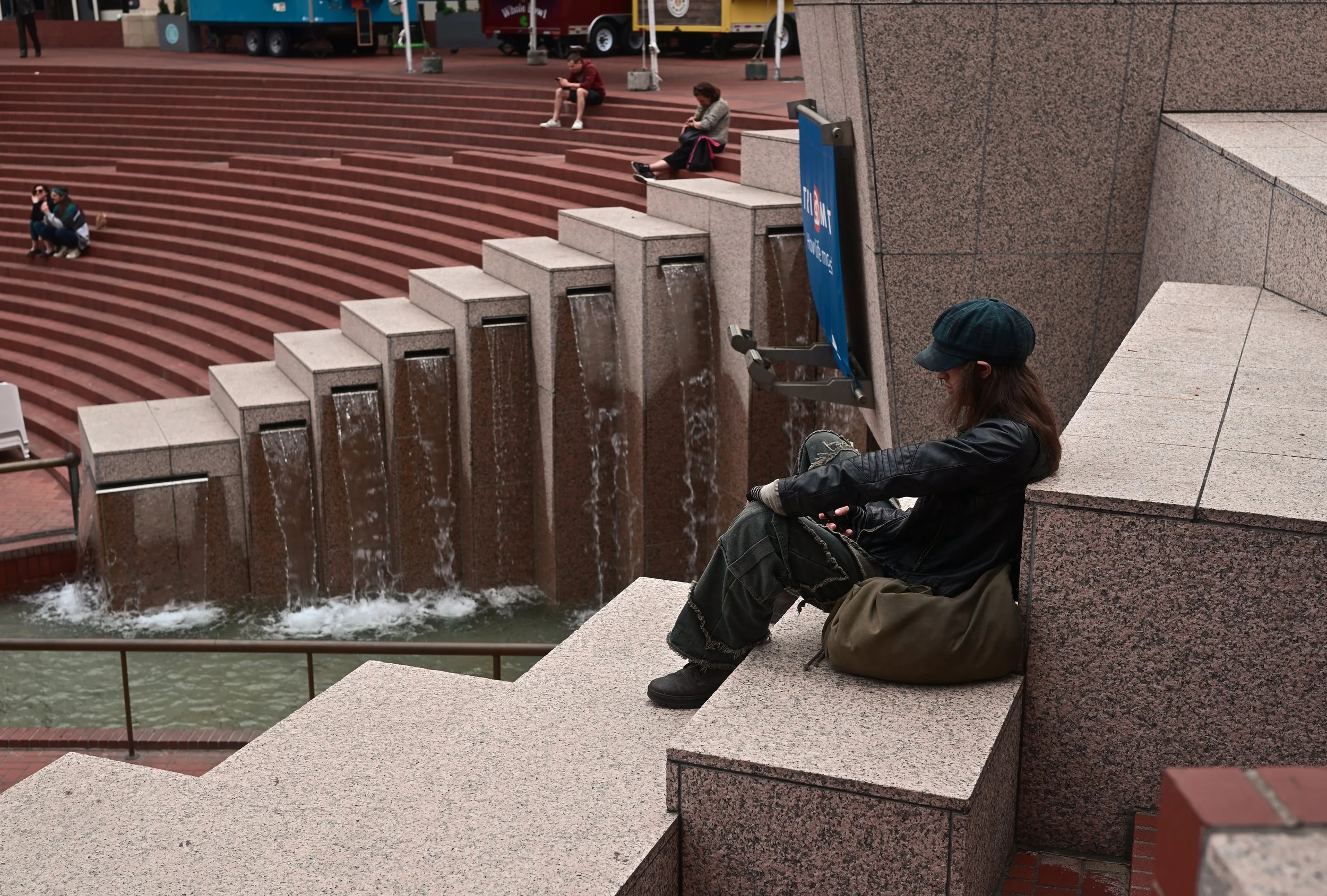 Several people sitting on stairs and ledges near a water feature, with some looking at their phones. The scene appears to be in an urban public space, with a water fountain and background vehicles.