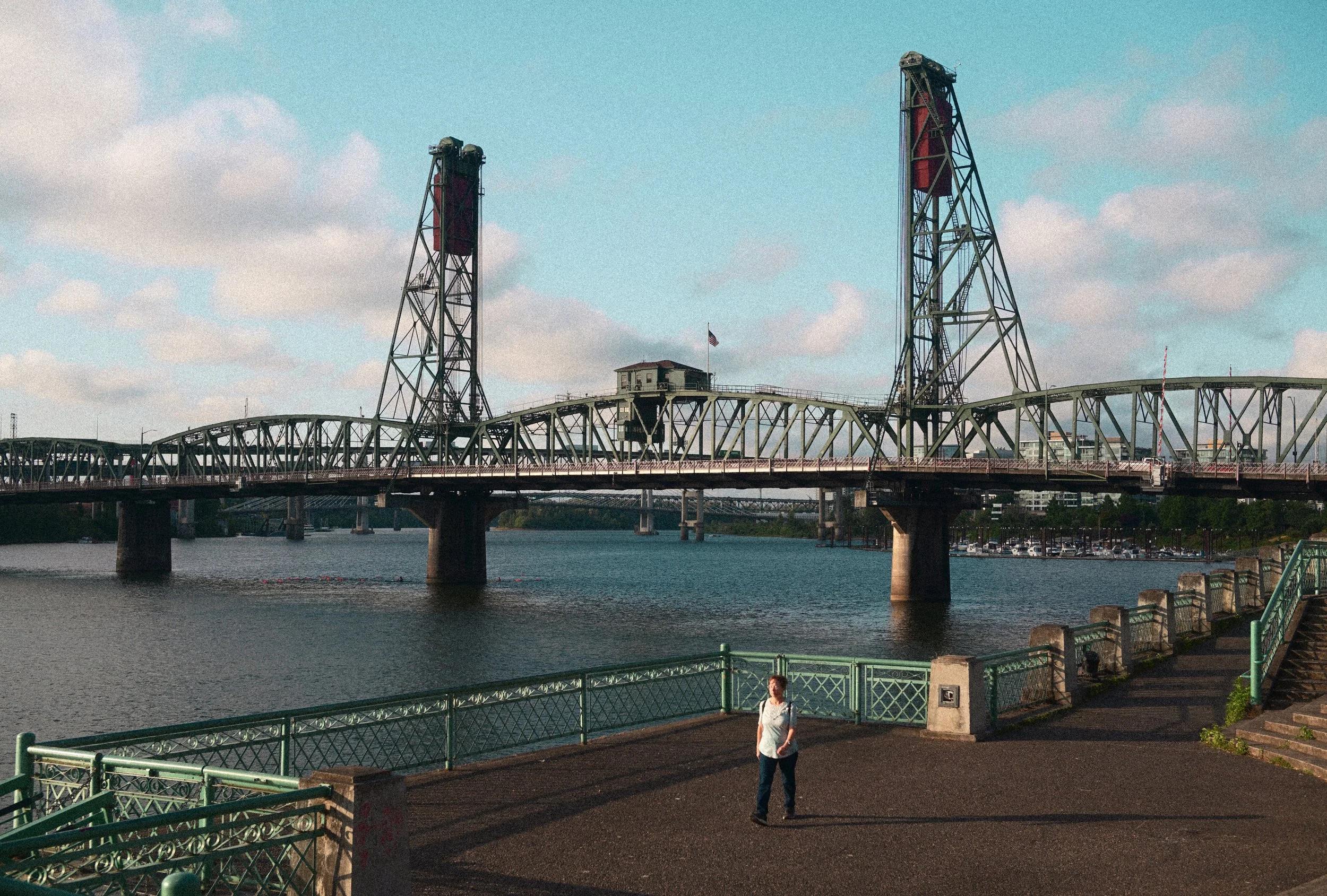 Historic steel lift bridge spanning a river, viewed from a waterfront promenade under a partly cloudy sky