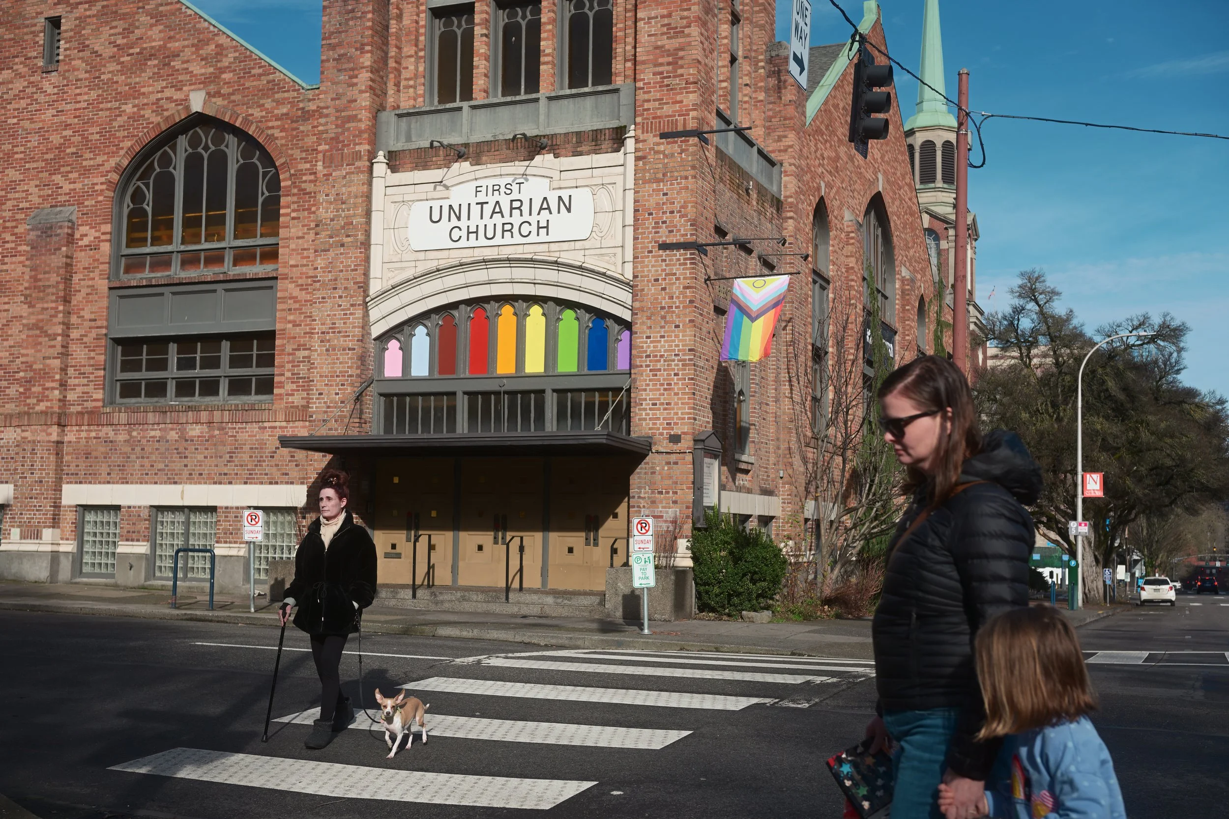 People crossing the street in front of a brick church with a rainbow pride flag displayed outside.