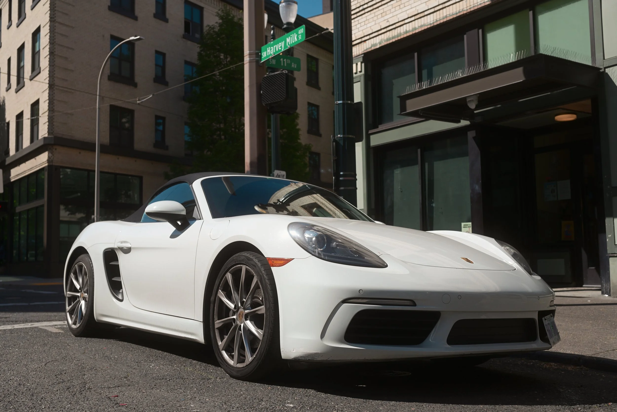 A white Porsche convertible sports car parked on the street at the corner of SW 11th Avenue and SW Harvey Milk Street in an urban area with buildings and street signs.