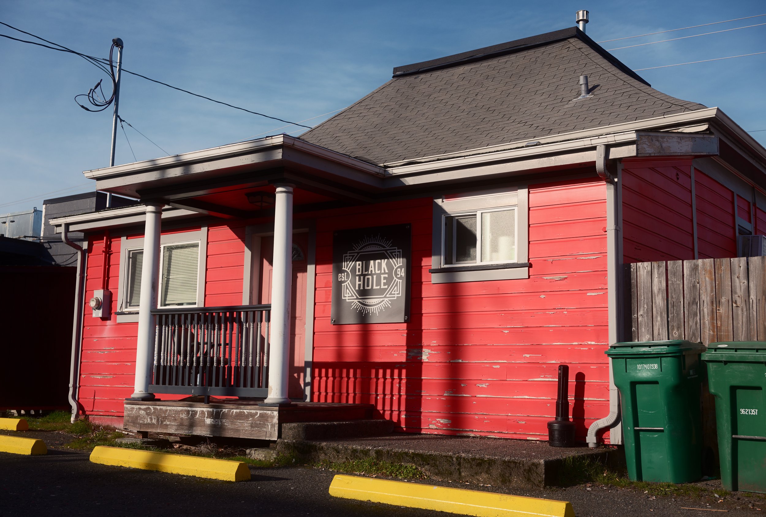 A two-story pink house with white trim and a small porch, with a sign on the front that reads "Black Hole Est. 94." The house has a gray shingle roof and a chimney, with outdoor electrical wires and green trash bins in front.