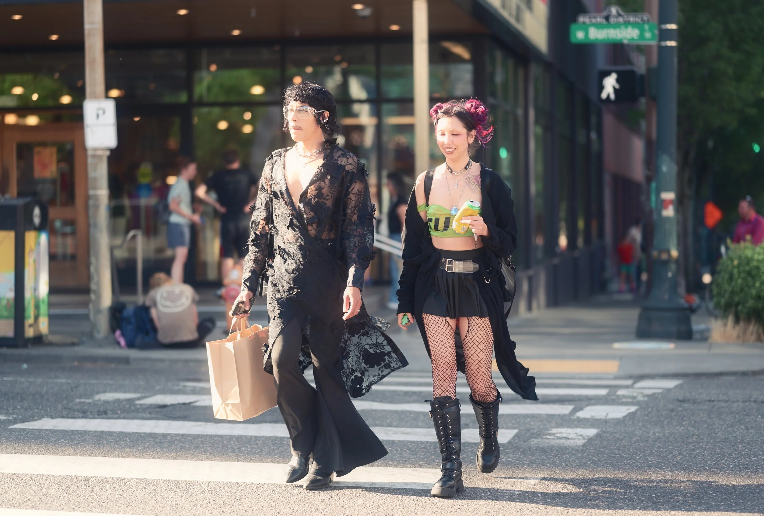 Two women walking across a pedestrian crosswalk in an urban area. One woman wears a black lace dress and glasses, carrying shopping bags. The other woman has pink hair, fishnet stockings, and wears a green tube top, black skirt, and combat boots, hol