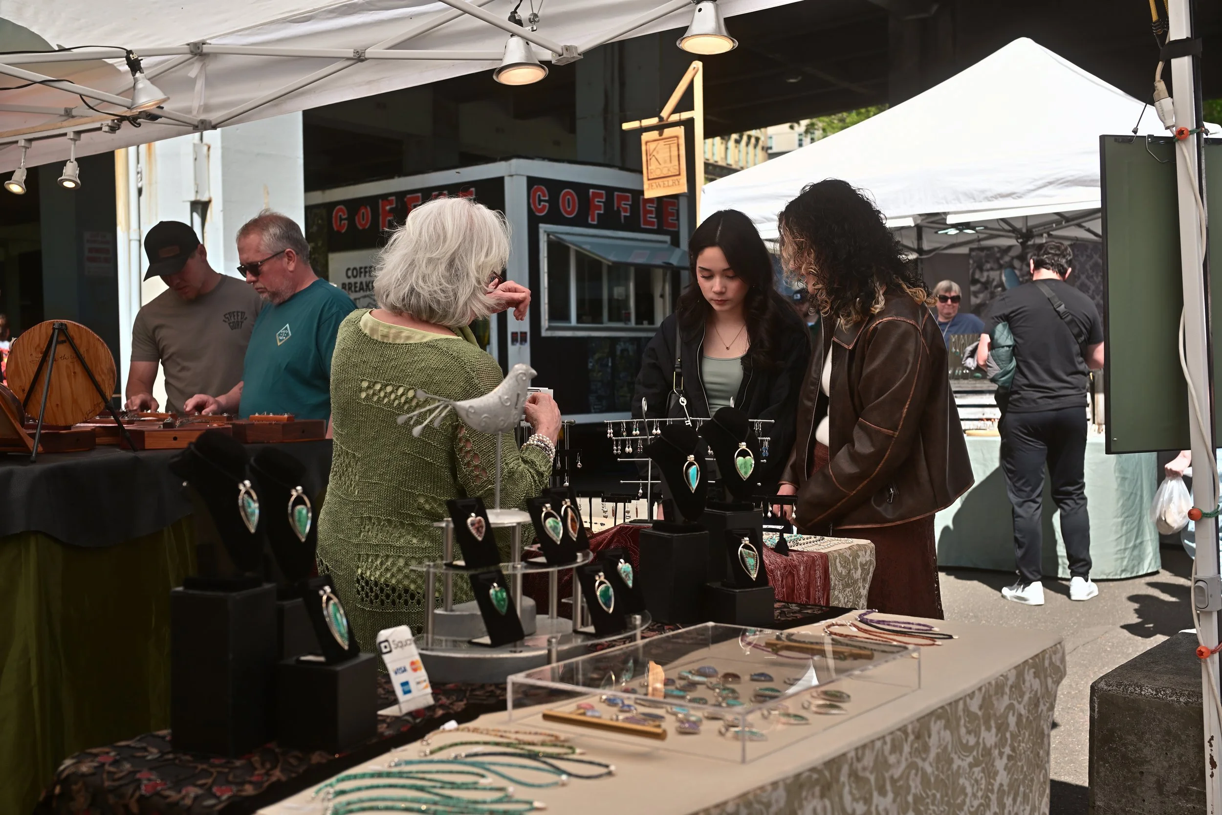 People shopping at an outdoor jewelry market stall with various jewelry items displayed on tables and racks.