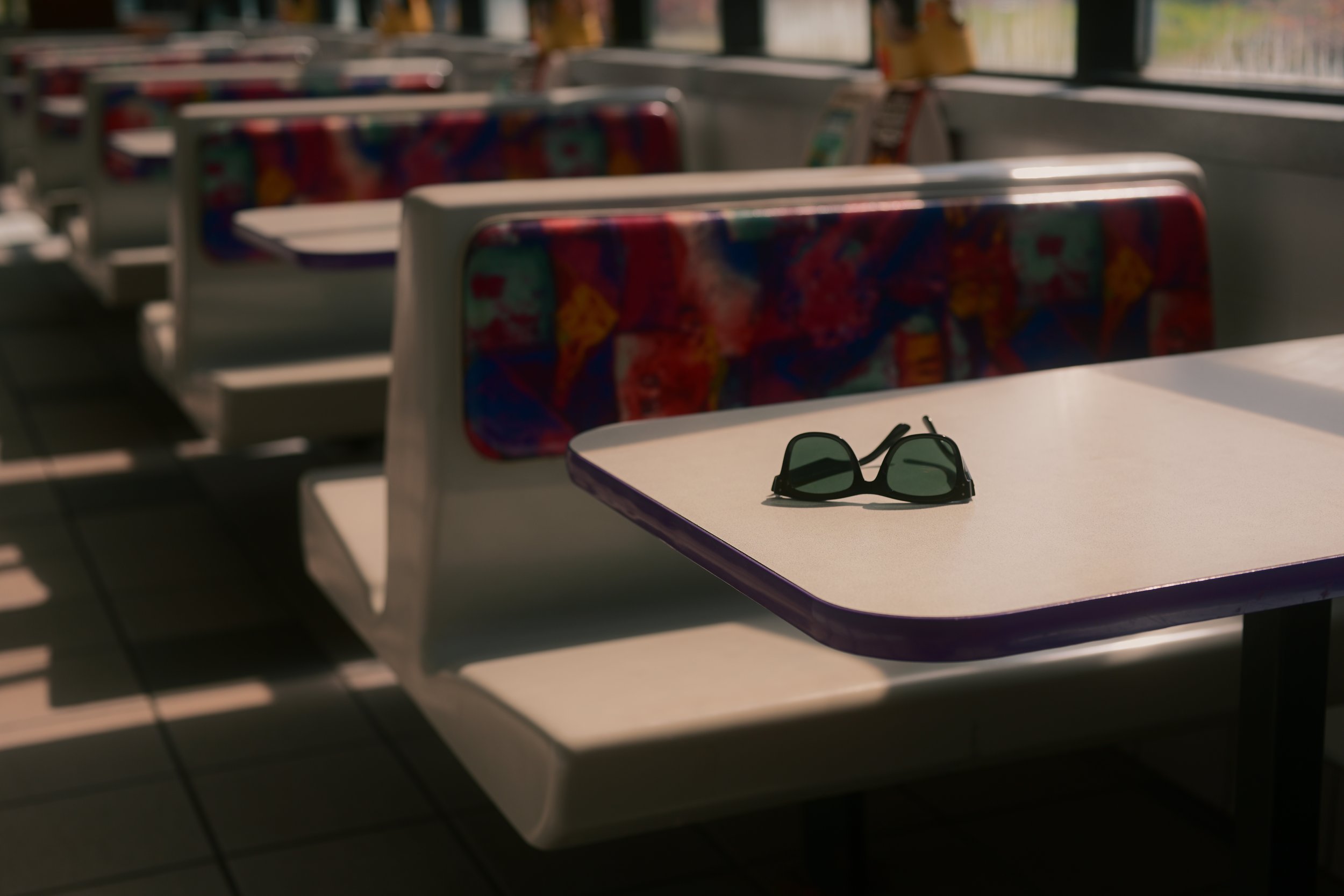 A pair of black sunglasses resting on a white table inside a bus or train. Rows of colorful patterned seats and windows are visible in the background.