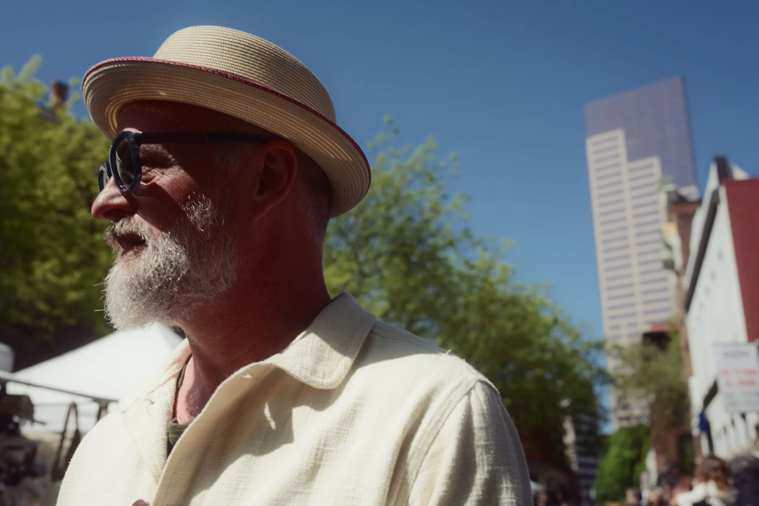 A man with a grey beard wearing a straw hat and sunglasses, standing outdoors under a blue sky with green trees and buildings in the background.