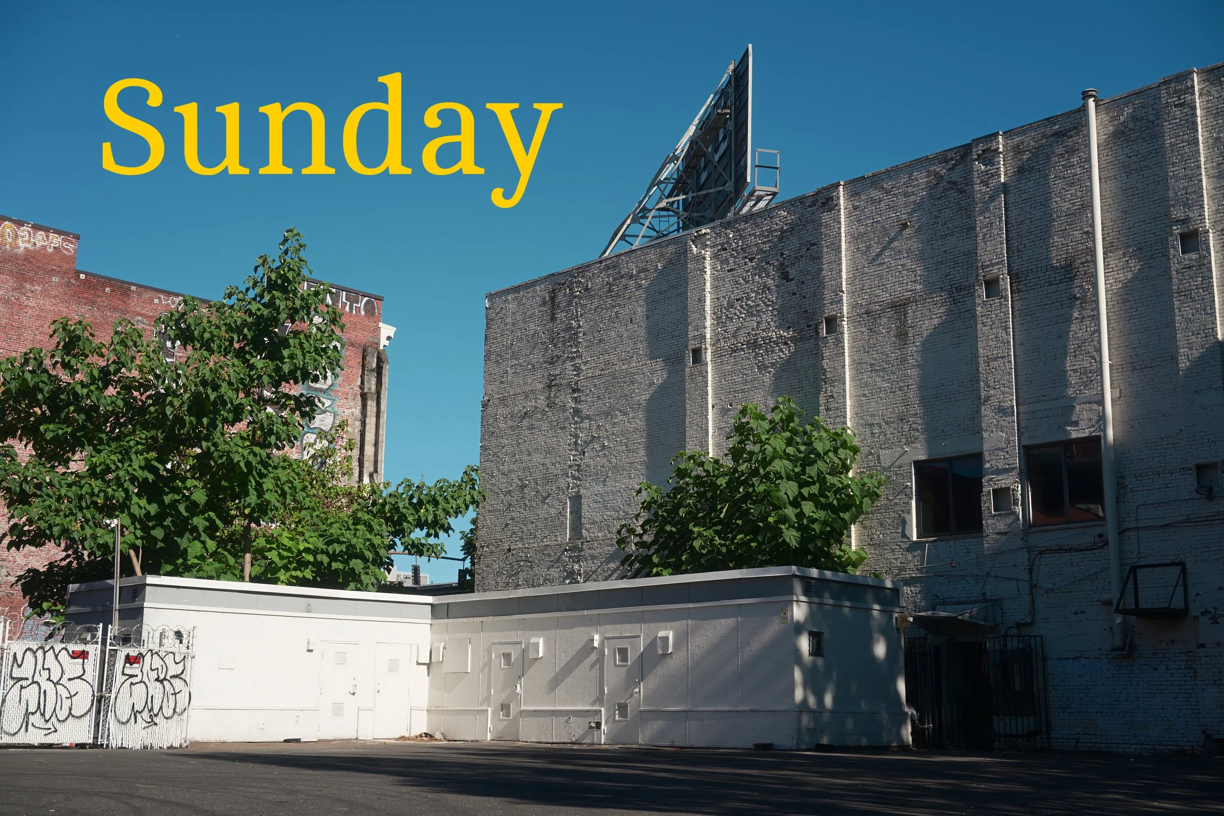 Urban scene with a large brick building, a billboard framework on the roof, trees in front, and a white mural on a fence at street level, under a clear blue sky.