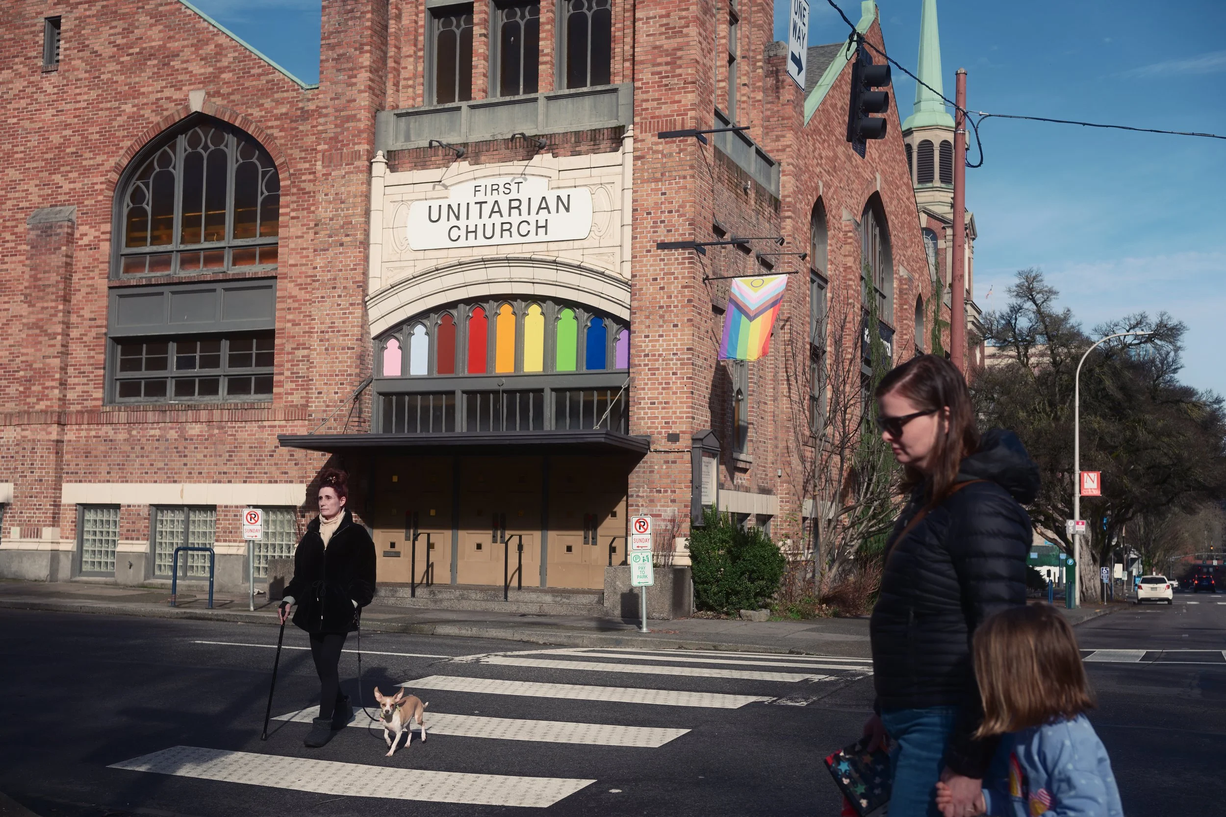 People cross the street in front of a brick church with a sign that reads 'First Unitarian Church' and rainbow flags, one of which has a rainbow pride flag. The scene is set on a sunny day with a blue sky.