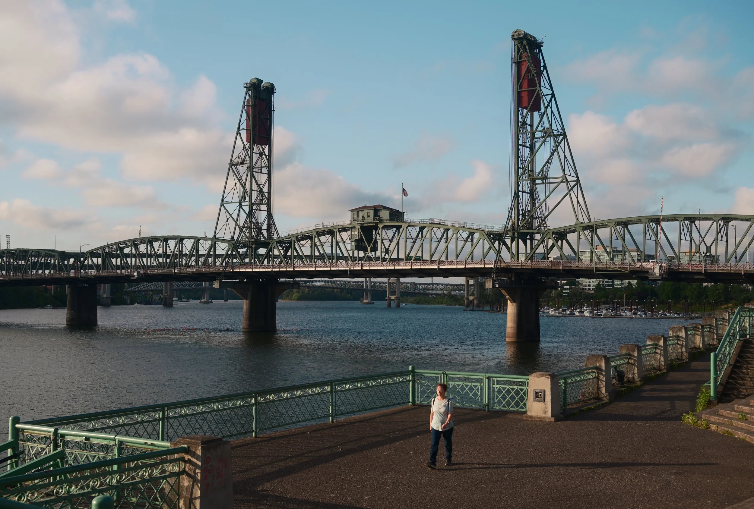 View of a bridge over a body of water with a small building and American flag atop on the bridge. A woman is walking along a paved path in the foreground, with a green railing beside the water.